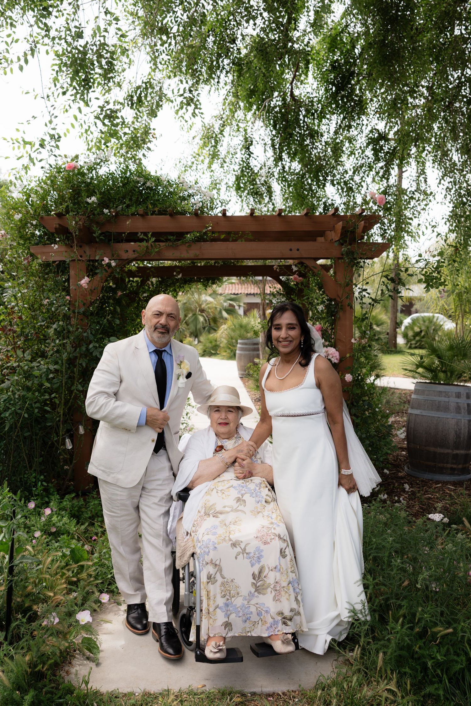 Bride and groom pose with elderly guest in wheelchair under wooden pergola in garden setting.