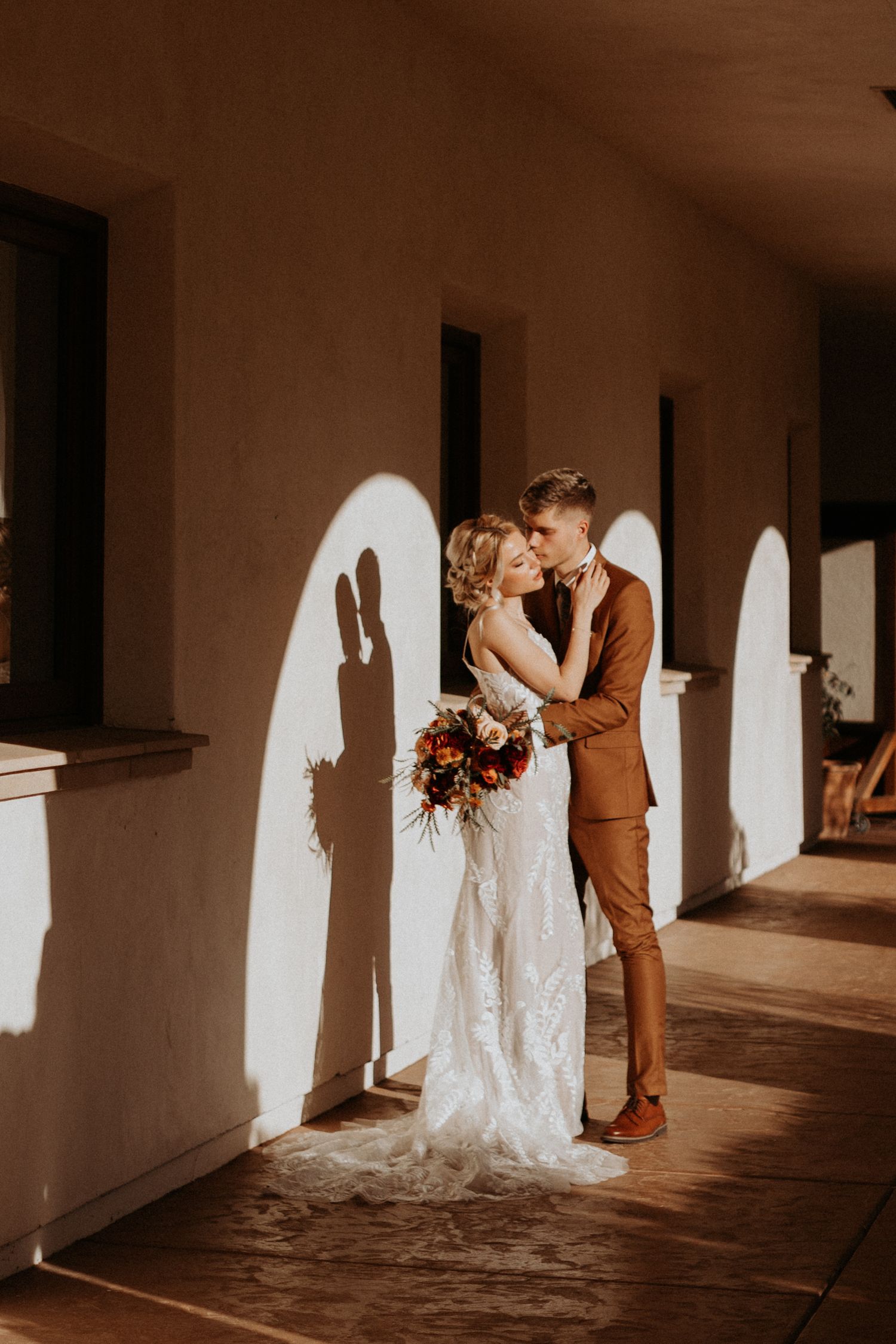 Bride and groom kissing in arched corridor with dramatic shadows cast on warm adobe walls.