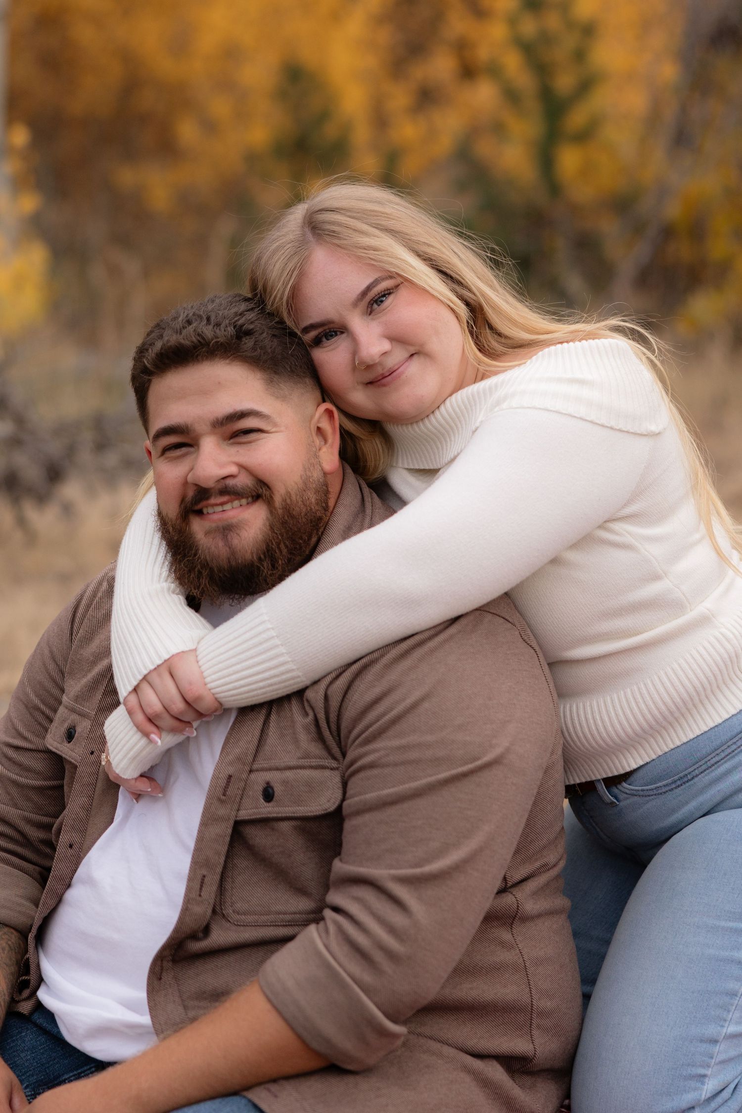 A smiling couple poses outdoors in autumn, woman in cream sweater hugging man in brown jacket with colorful fall foliage behind.