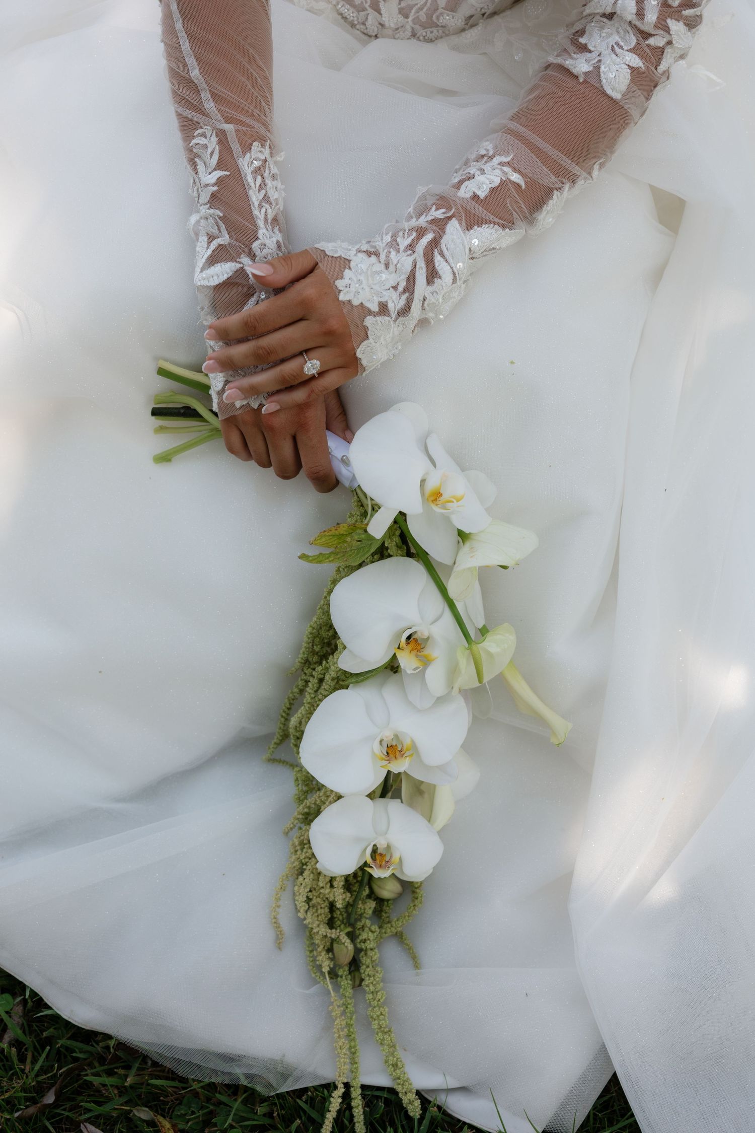 Bride in lace wedding gown holding elegant white orchid bouquet with greenery accents on her lap.