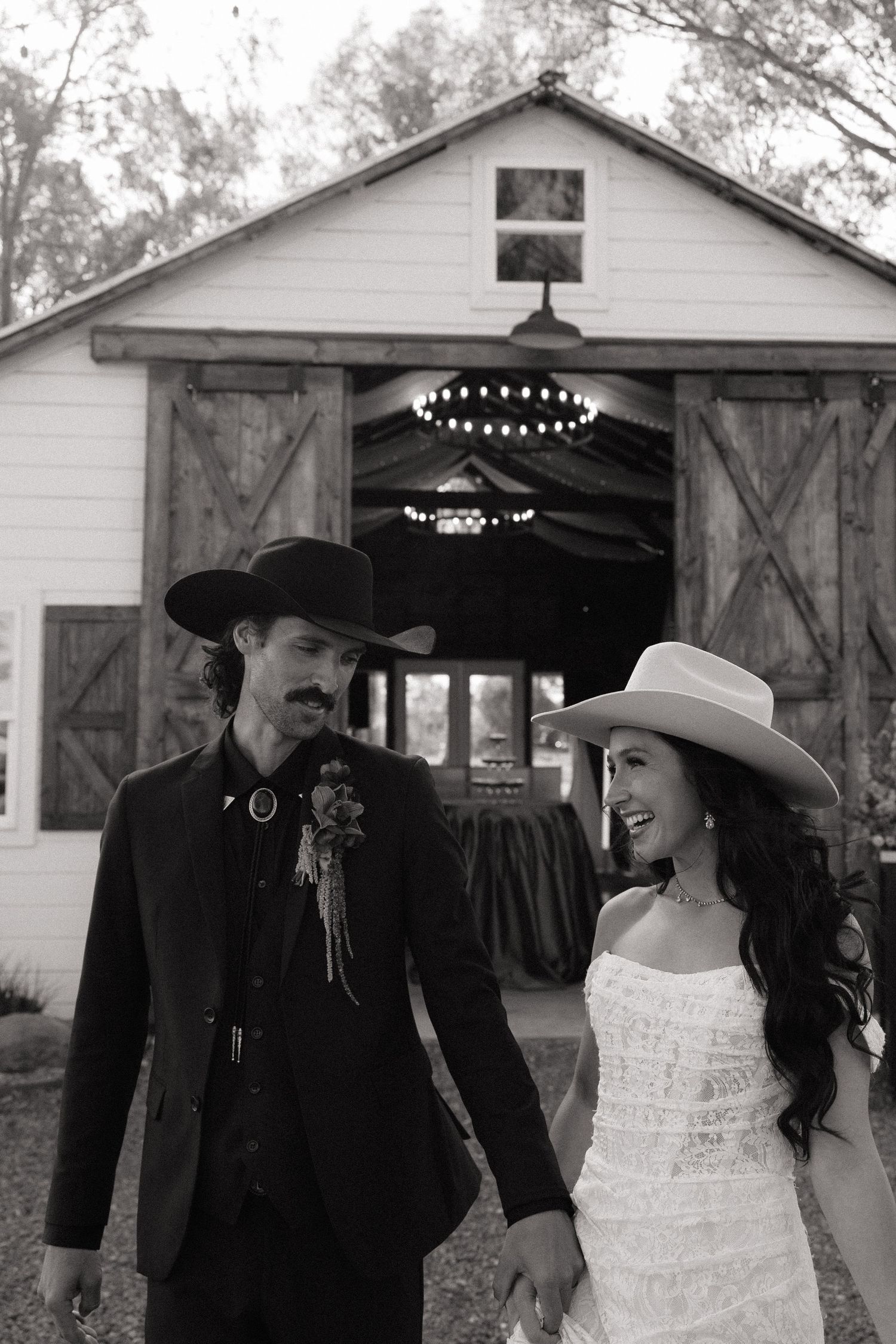 Black and white photo of bride and groom in cowboy hats walking hand in hand outside a barn.