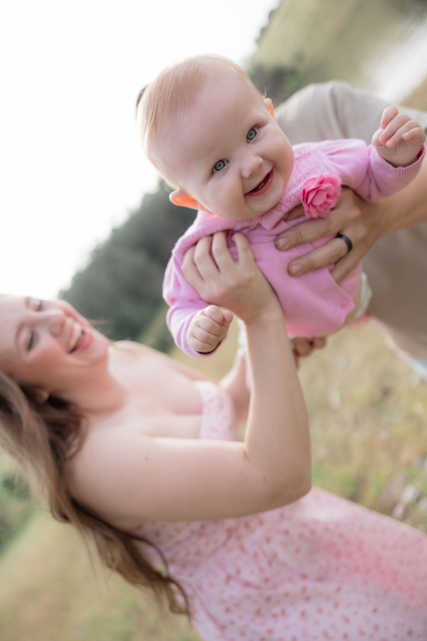Mother joyfully lifting smiling baby girl in pink outfit outdoors in nature.