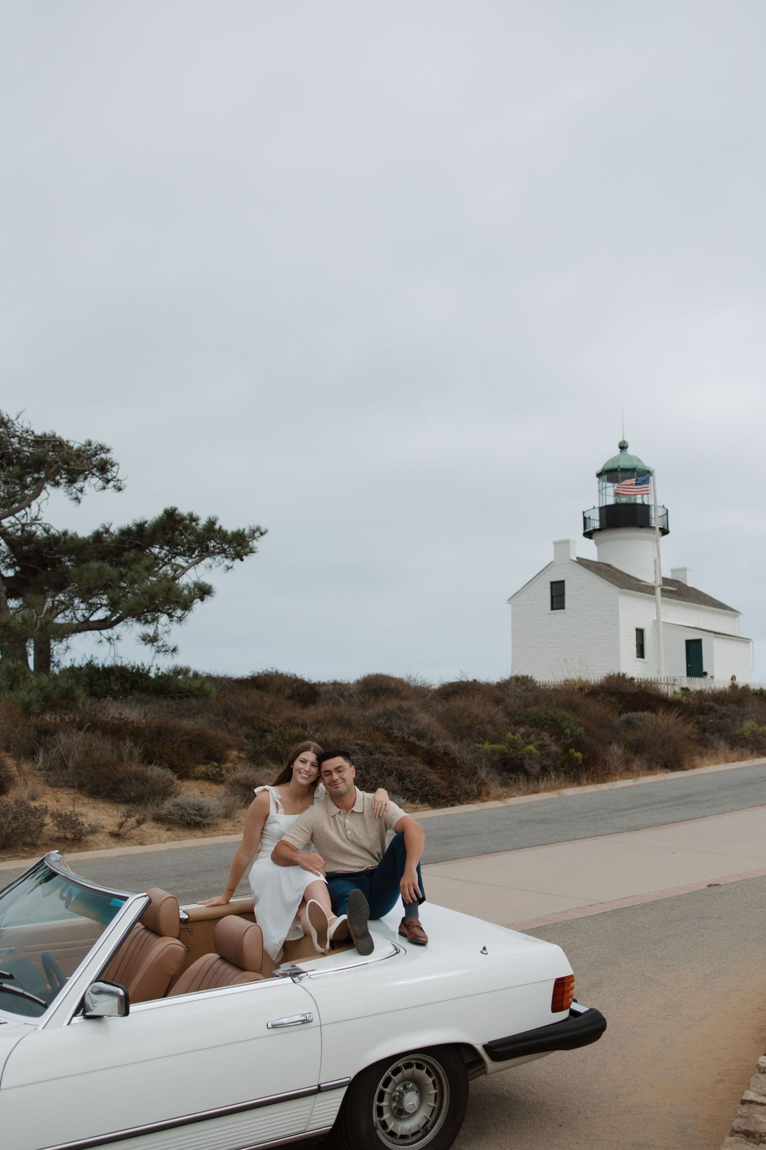 Couple posing on a white convertible car with a lighthouse in the background.