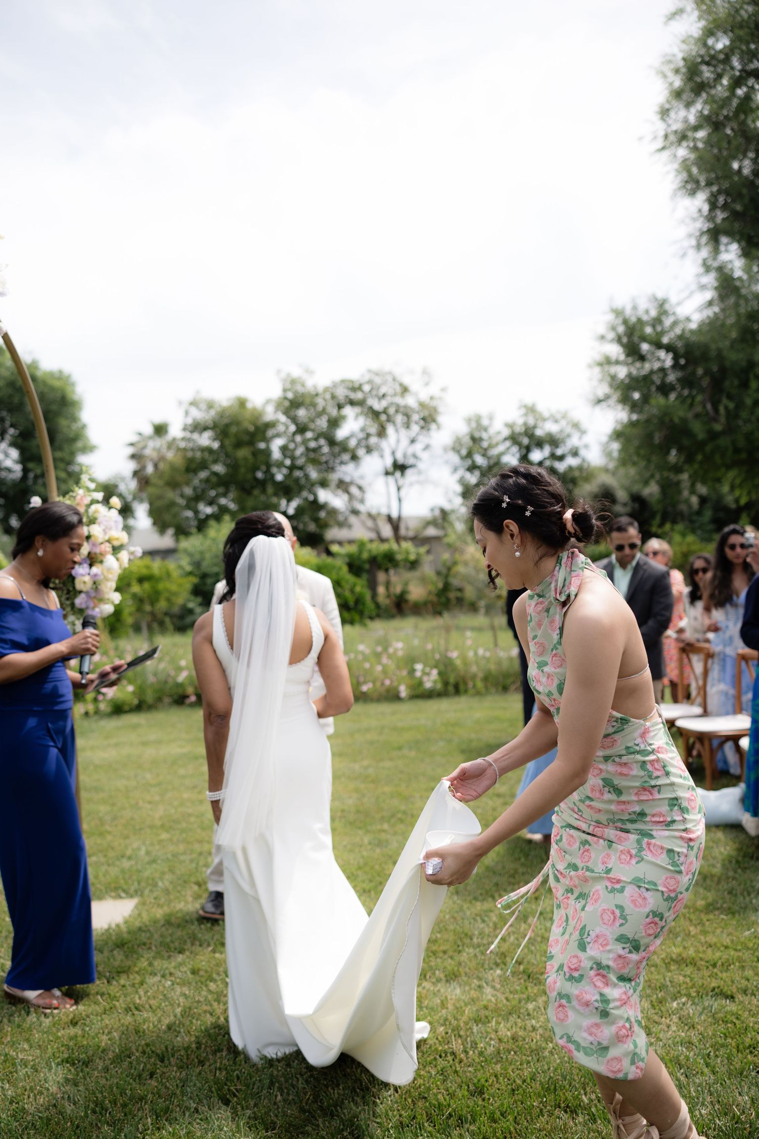 Bridesmaid in floral dress adjusts bride's white wedding gown train during outdoor garden ceremony.