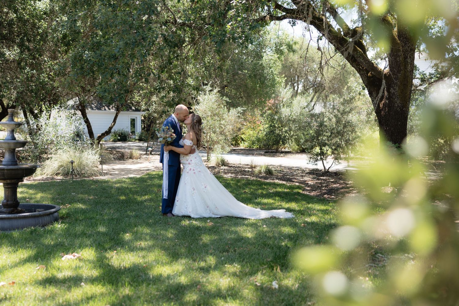 Bride and groom sharing a romantic kiss in a lush garden with fountain and olive trees.