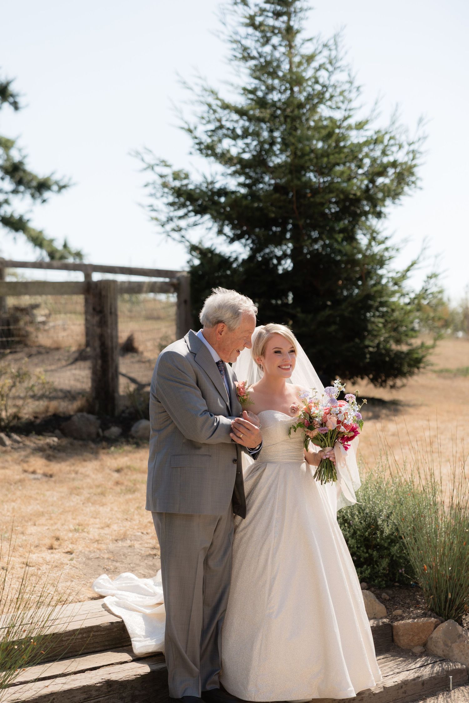 Bride in white gown holds pink bouquet beside older man in gray suit outdoors near pine tree on sunny wedding day.