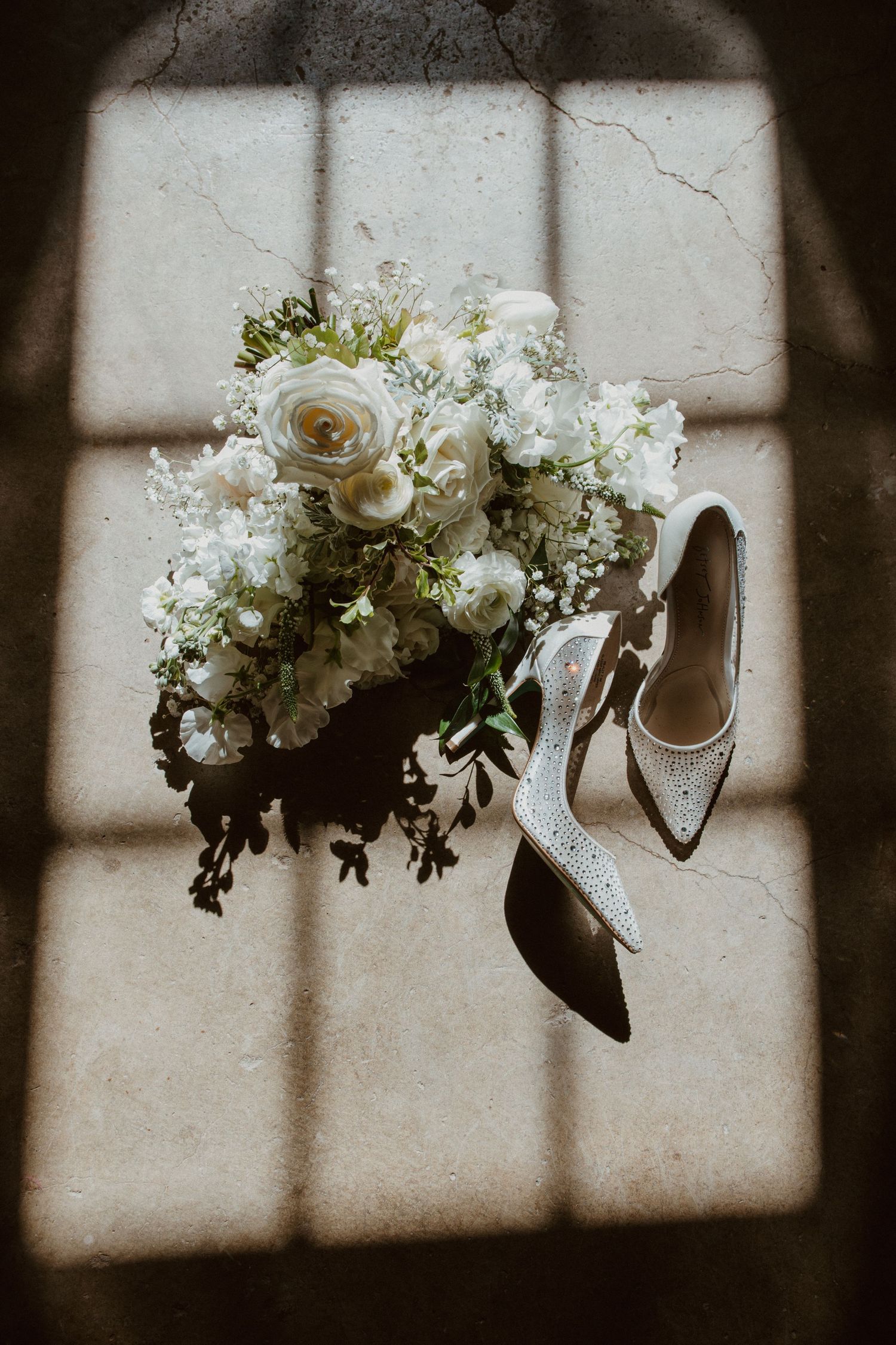 White wedding bouquet with roses beside sparkly bridal heels on textured floor with shadow pattern.