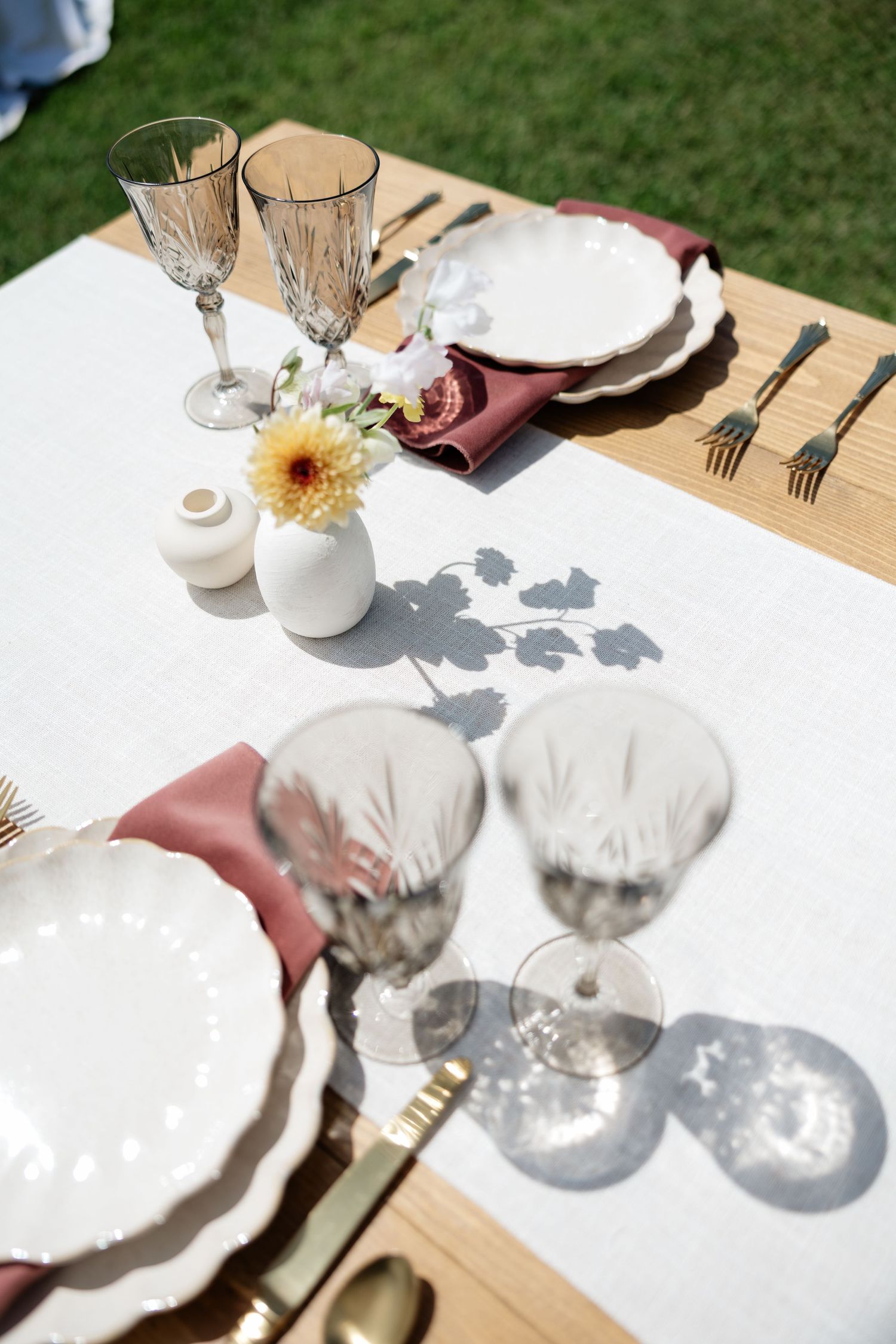 Elegant outdoor wedding table setting with crystal glasses, white plates, gold cutlery, and fresh flowers in sunlight.
