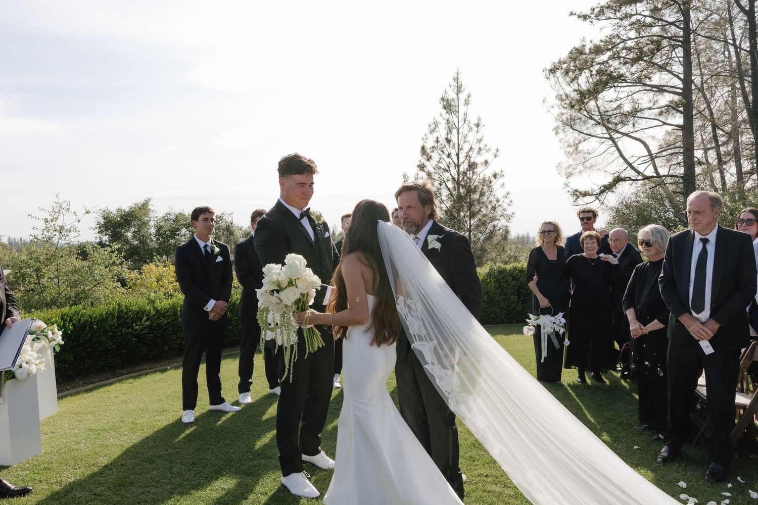 Bride and groom exchanging vows at outdoor wedding ceremony with guests standing.