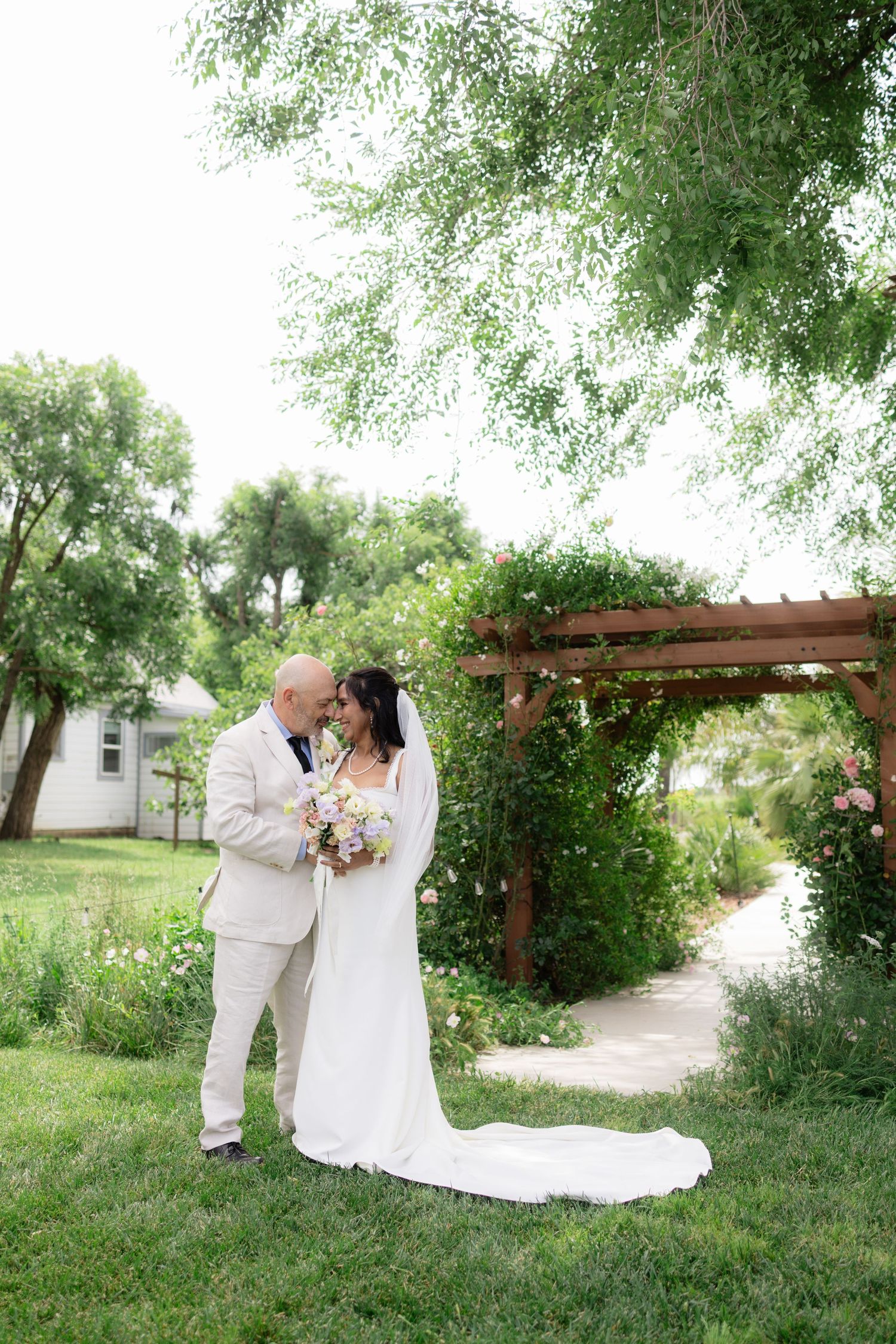 Bride and groom sharing romantic moment under wooden pergola surrounded by lush greenery at garden wedding.