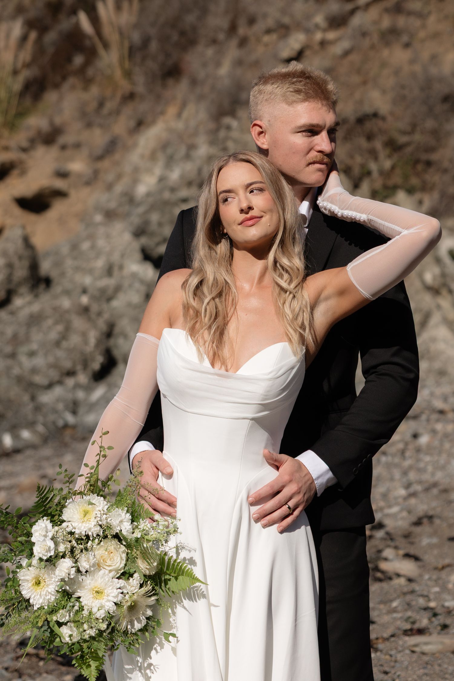 Bride in white off-shoulder gown holds bouquet while groom in black tux embraces her against rocky backdrop.