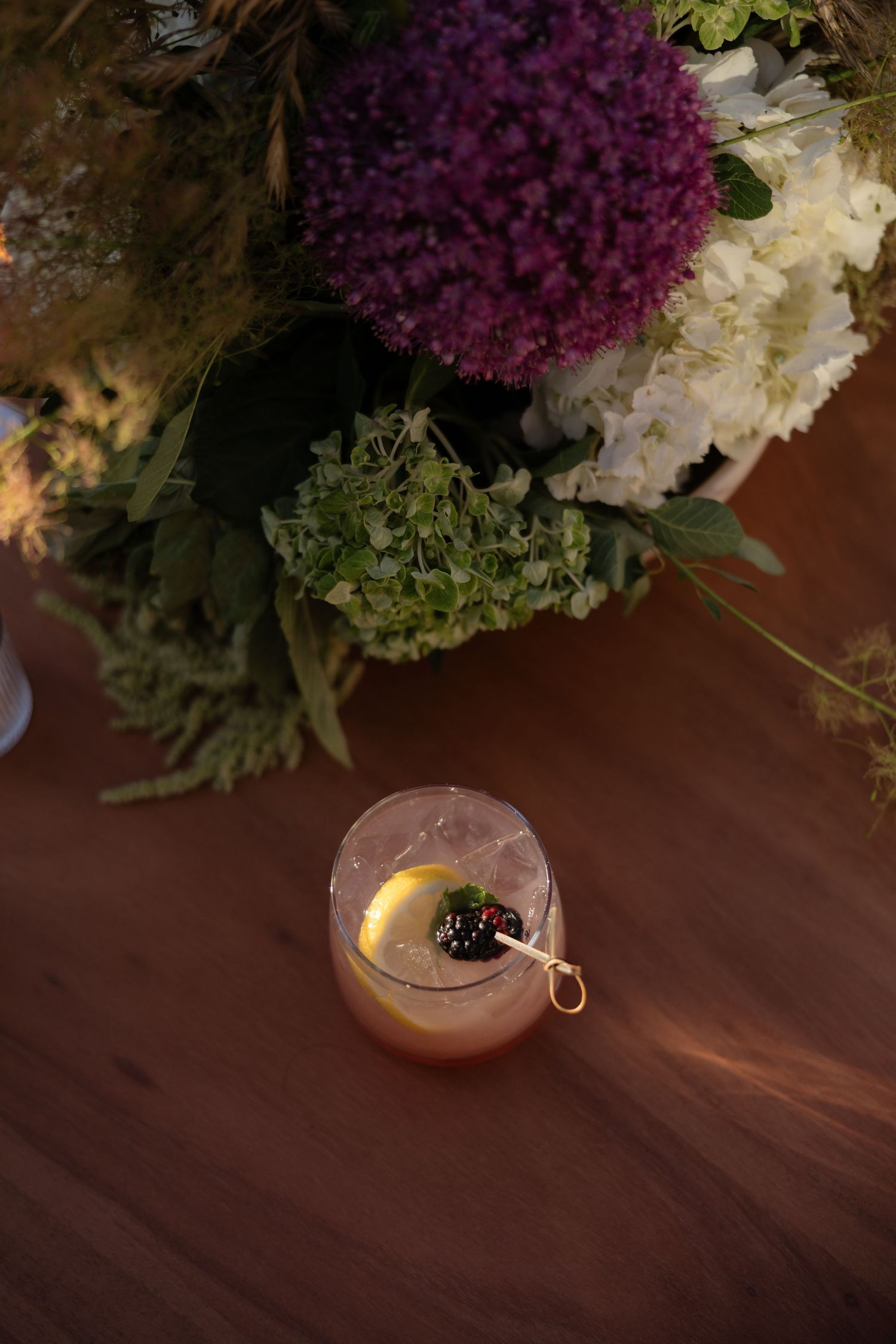 A pink cocktail garnished with lemon and blackberry sits on a wood table beside a lush floral arrangement.