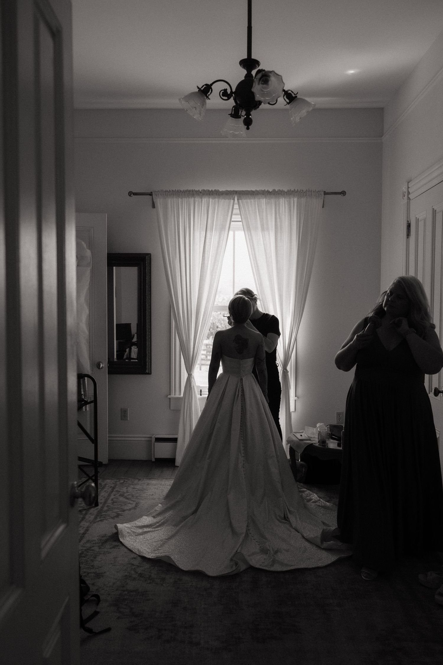 Black and white silhouette of a bride in a ball gown wedding dress standing before a window with flowing curtains.