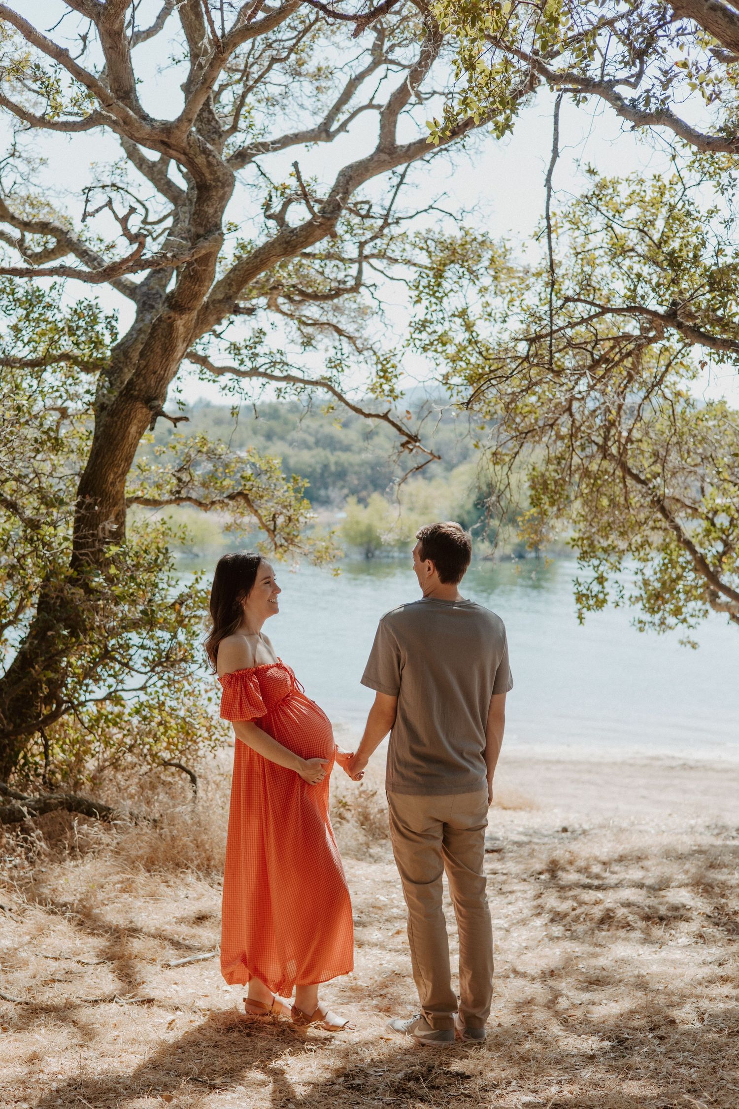 Pregnant woman in red dress holds hands with partner by a scenic lake under oak trees.