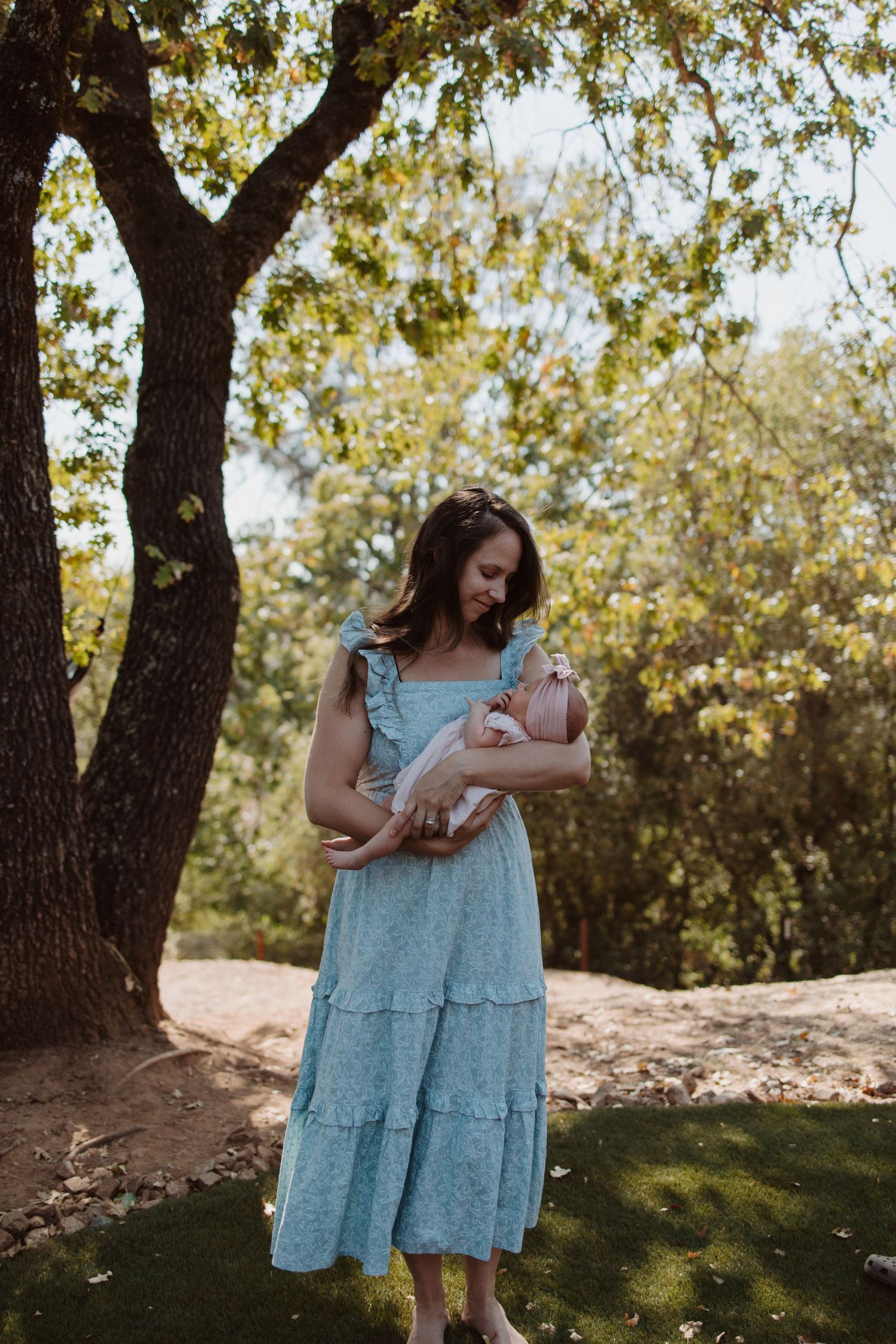 Mother in blue dress holds newborn baby outdoors under a large oak tree.