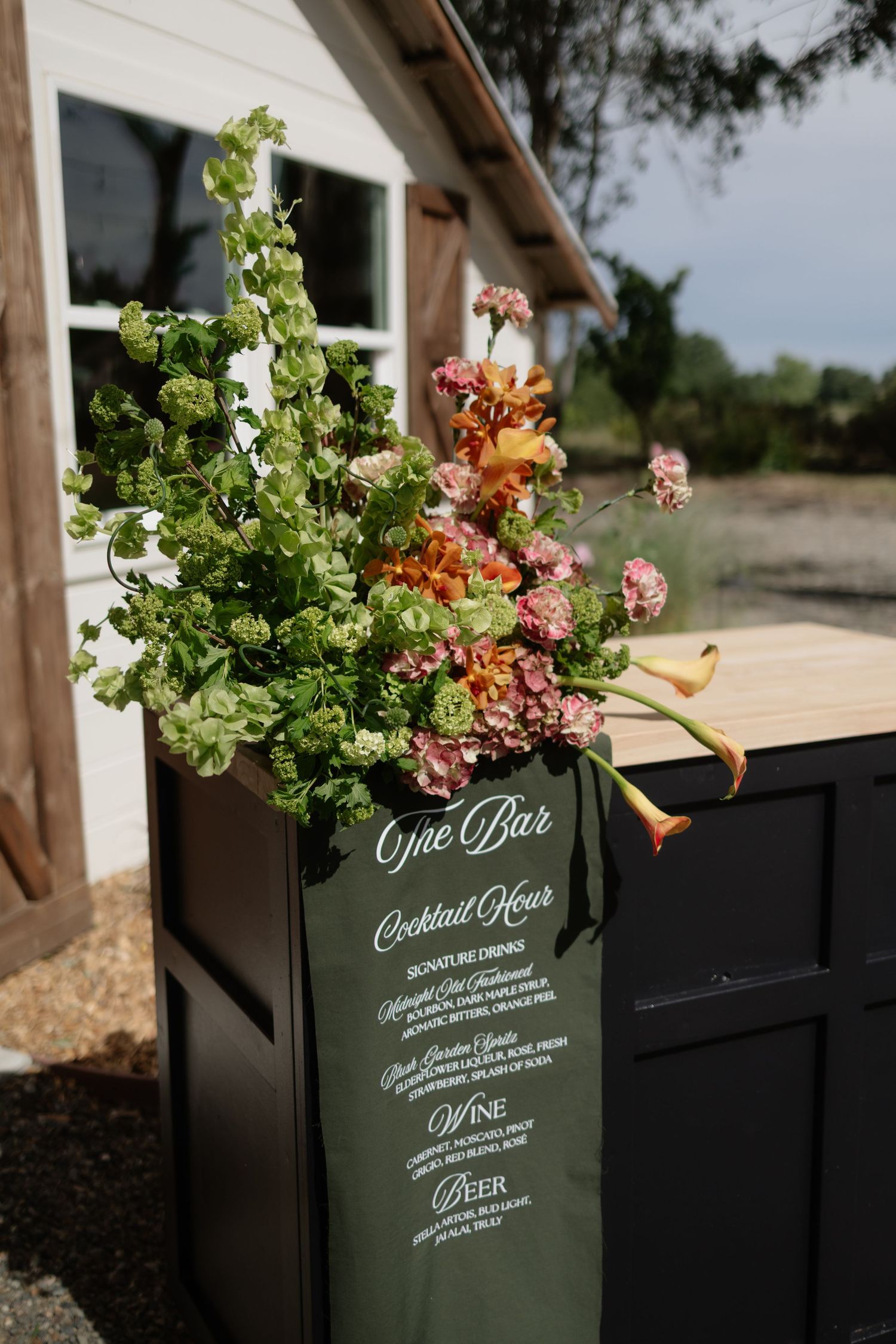 Elegant bar sign for cocktail hour with lush floral arrangement of green hops, pink hydrangeas, and orange calla lilies.