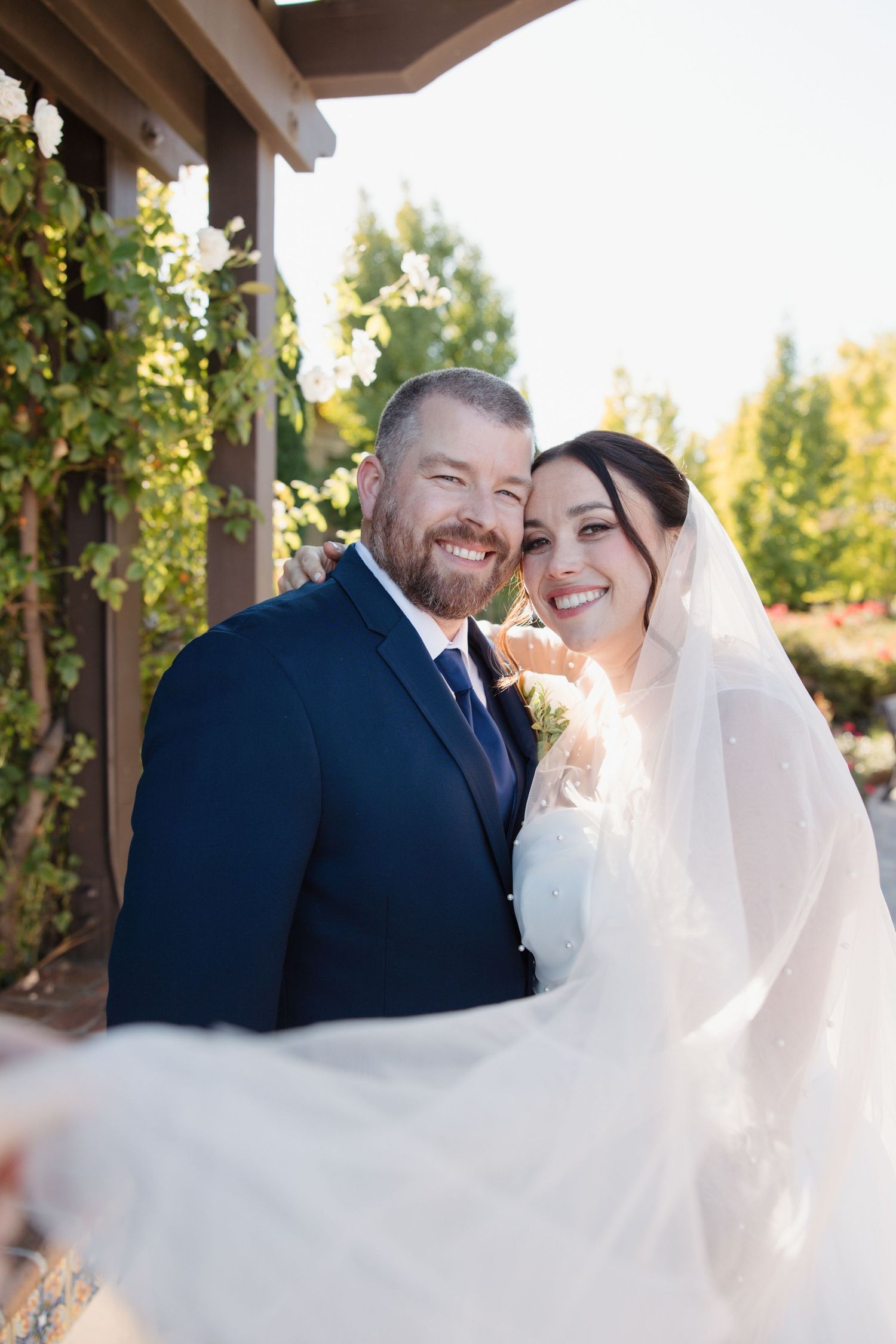 Smiling bride and groom pose together outdoors under a pergola on their wedding day.