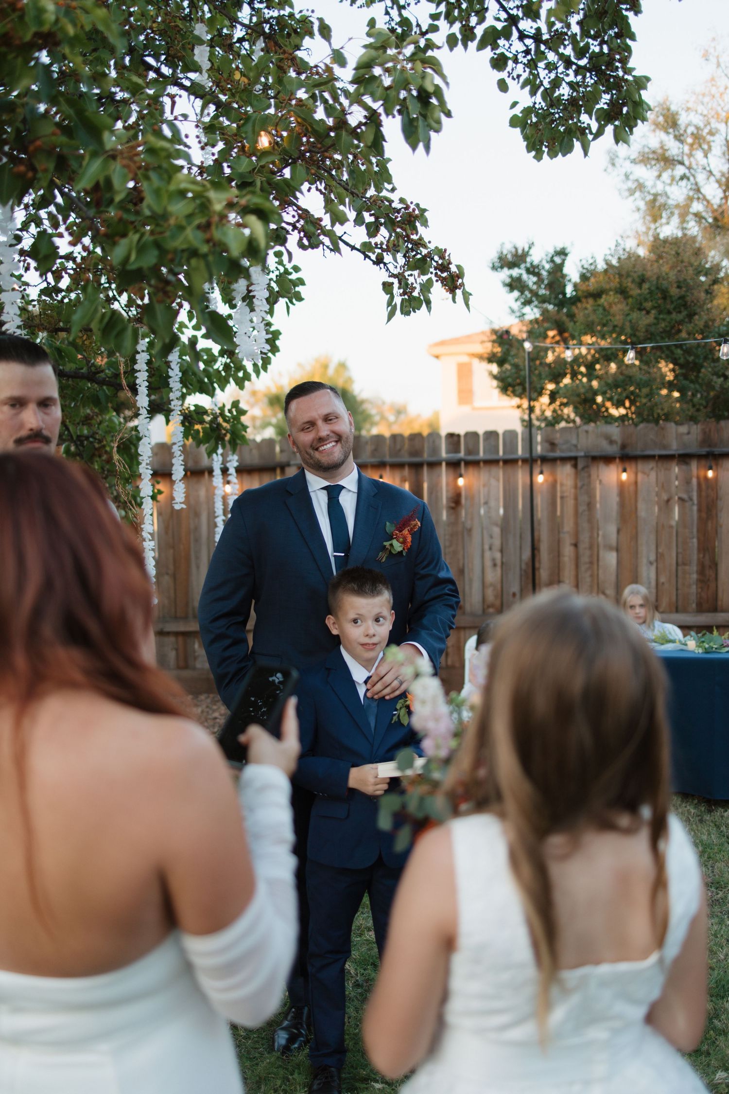 Smiling groom in navy suit stands with young boy at backyard wedding ceremony.