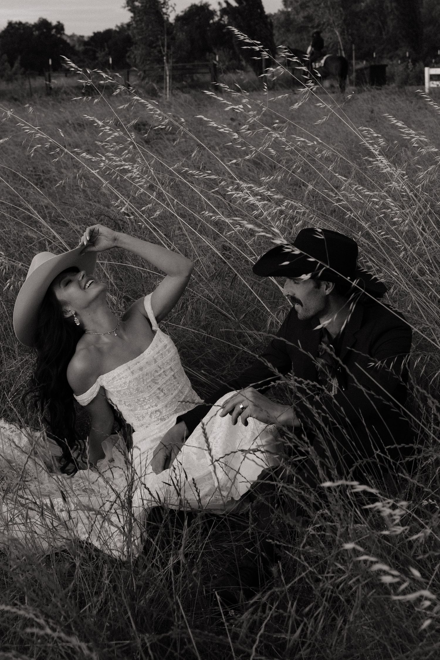 Bride and groom in cowboy hats relaxing together in tall grass in a romantic outdoor setting.