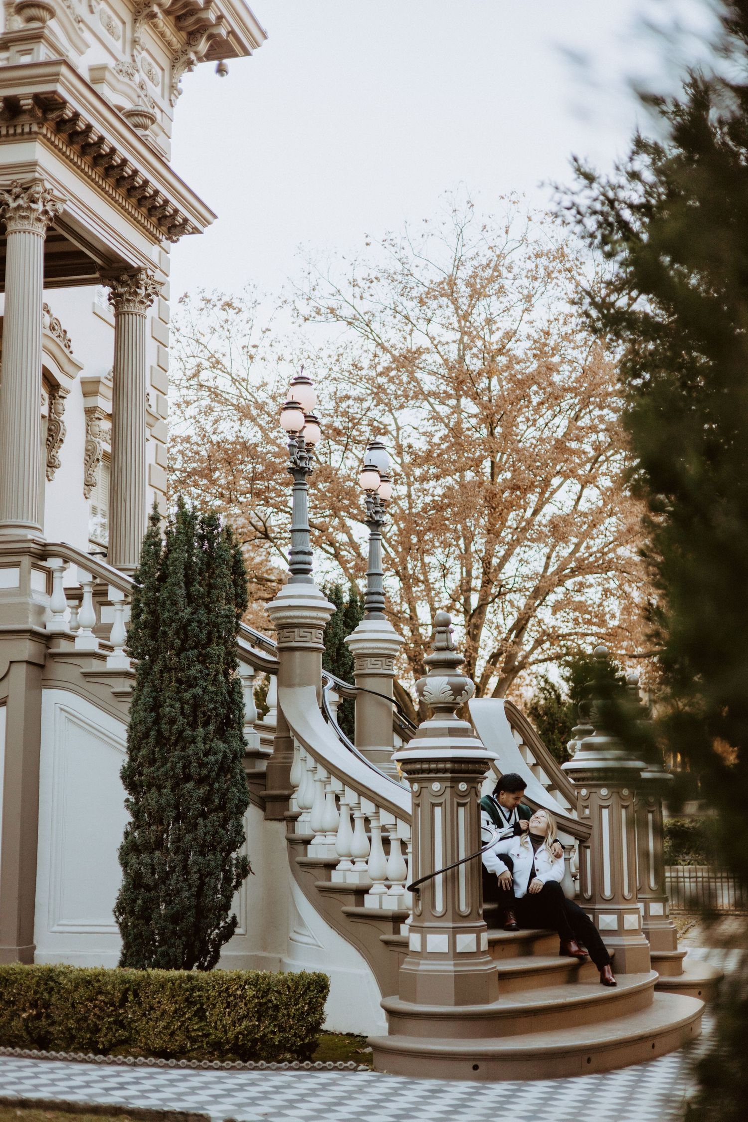 Couple sitting on ornate white staircase of elegant Victorian mansion surrounded by cypress trees.