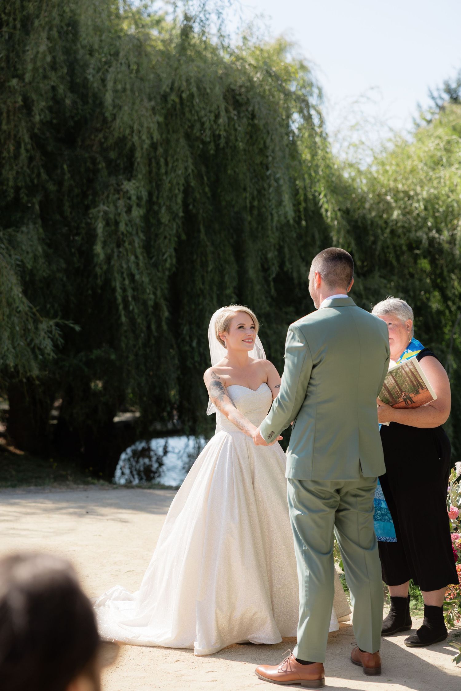 Bride and groom exchange vows outdoors under a weeping willow tree, officiant holding book beside them.