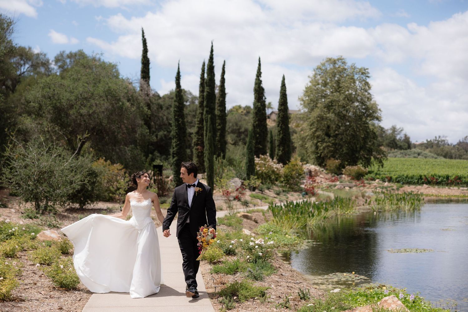 Bride and groom walk hand in hand along scenic pond path with cypress trees at wedding.