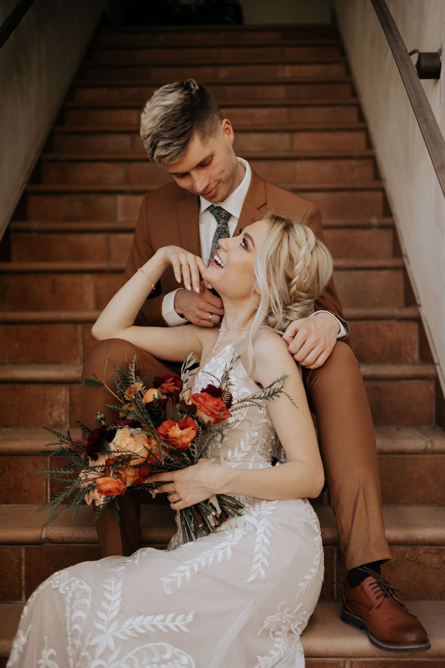 Bride and groom on rustic stairs, groom in brown suit, bride holding vibrant autumn bouquet.