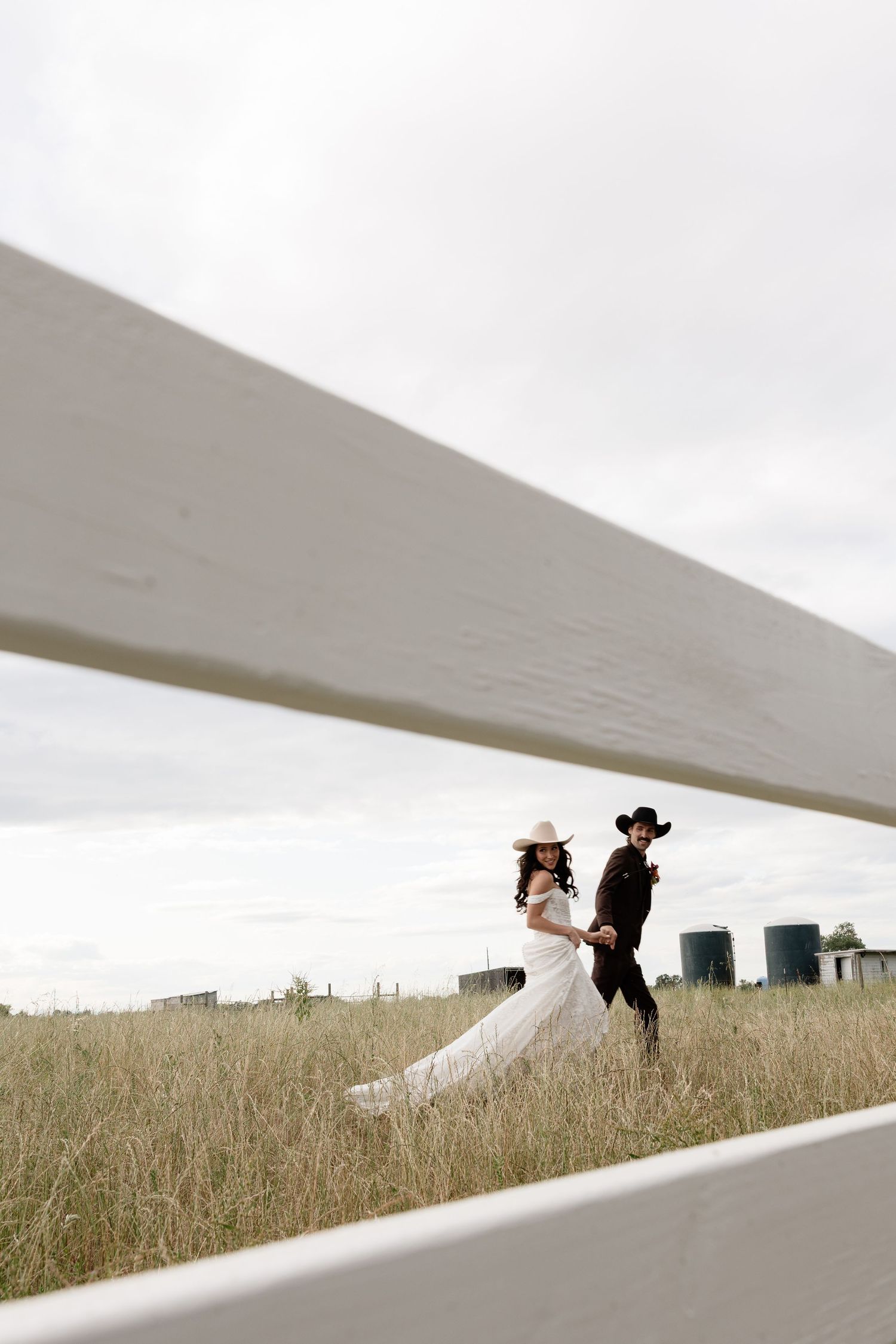 A bride in a white gown and groom in a black cowboy hat walk hand in hand through a grassy field.