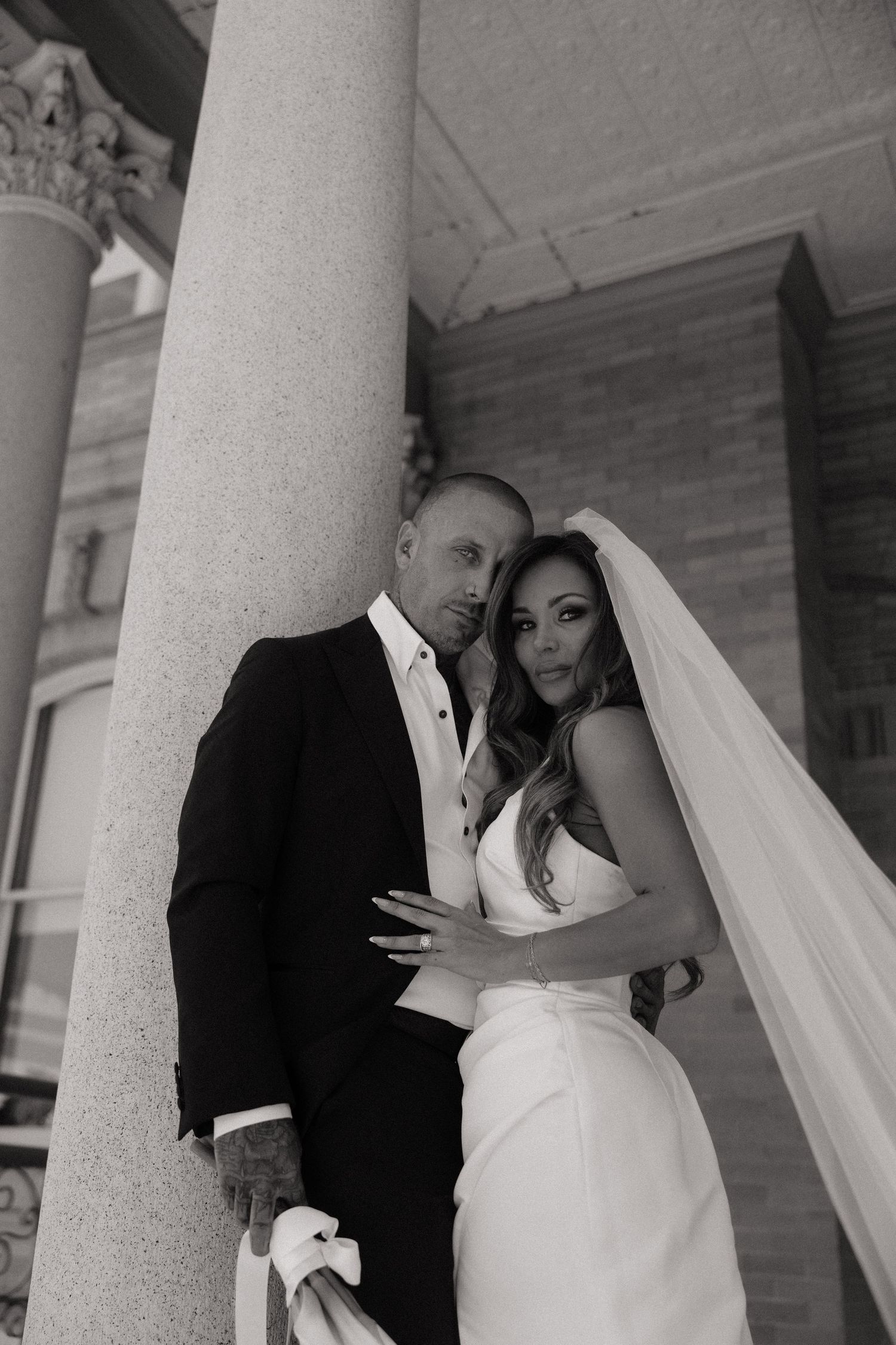 Black and white wedding photo of bride in white gown and veil with groom in black suit leaning against stone column.