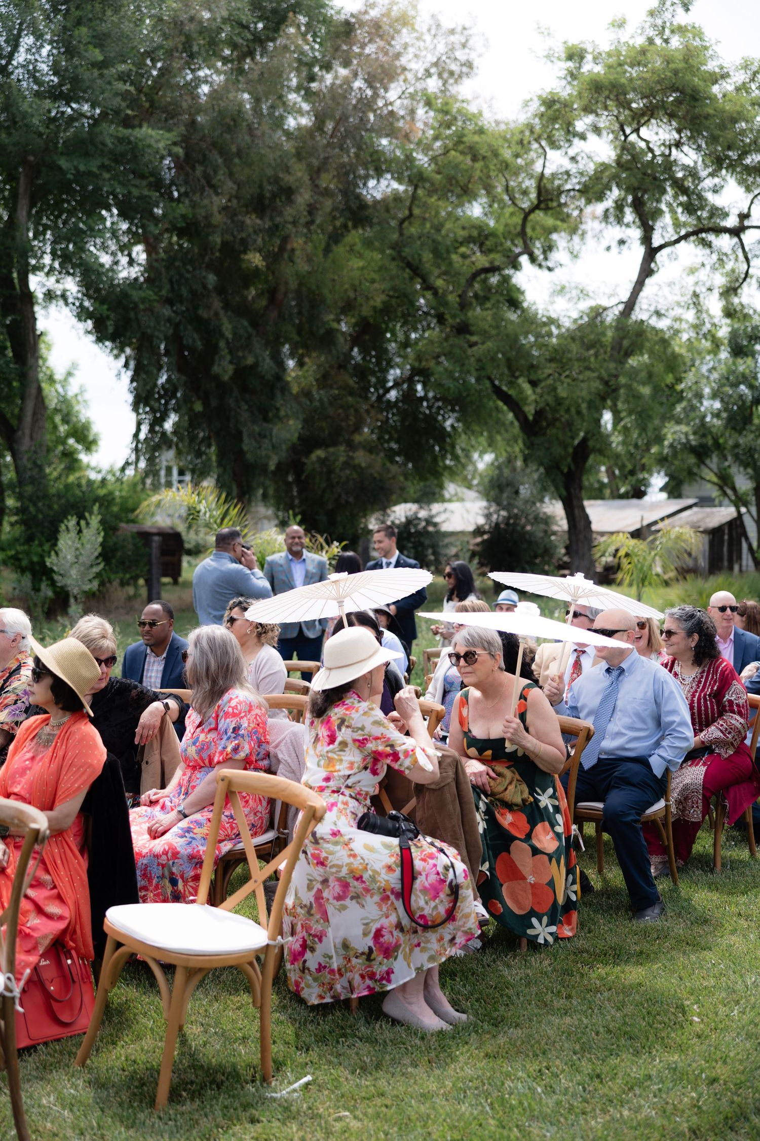 Wedding guests seated outdoors on wooden chairs under trees, some holding parasols for sun protection.