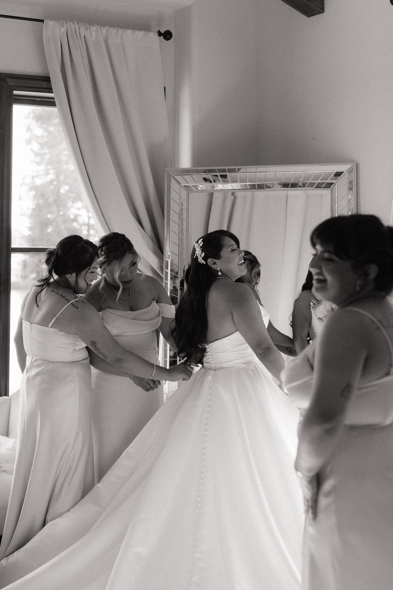 Black and white photo of a bride being helped into her wedding gown by bridesmaids near a mirror.