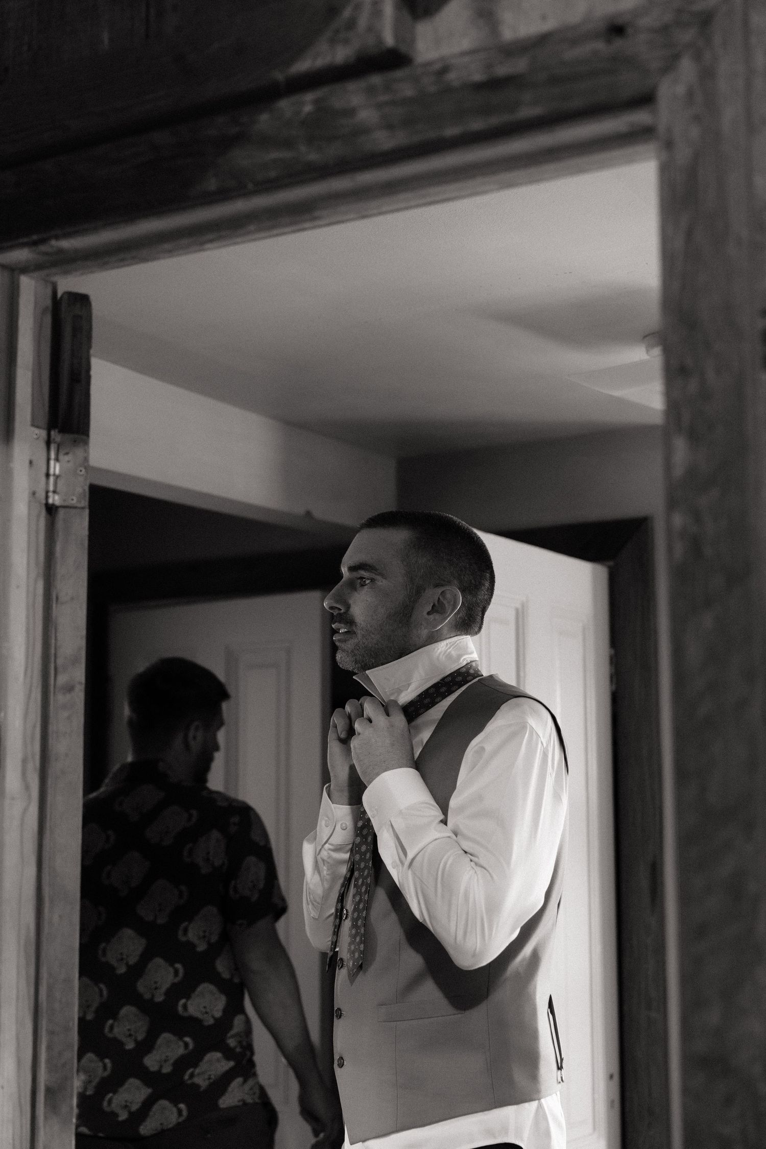 Black and white photo of a groom adjusting his tie in a doorway while getting ready for his wedding ceremony.