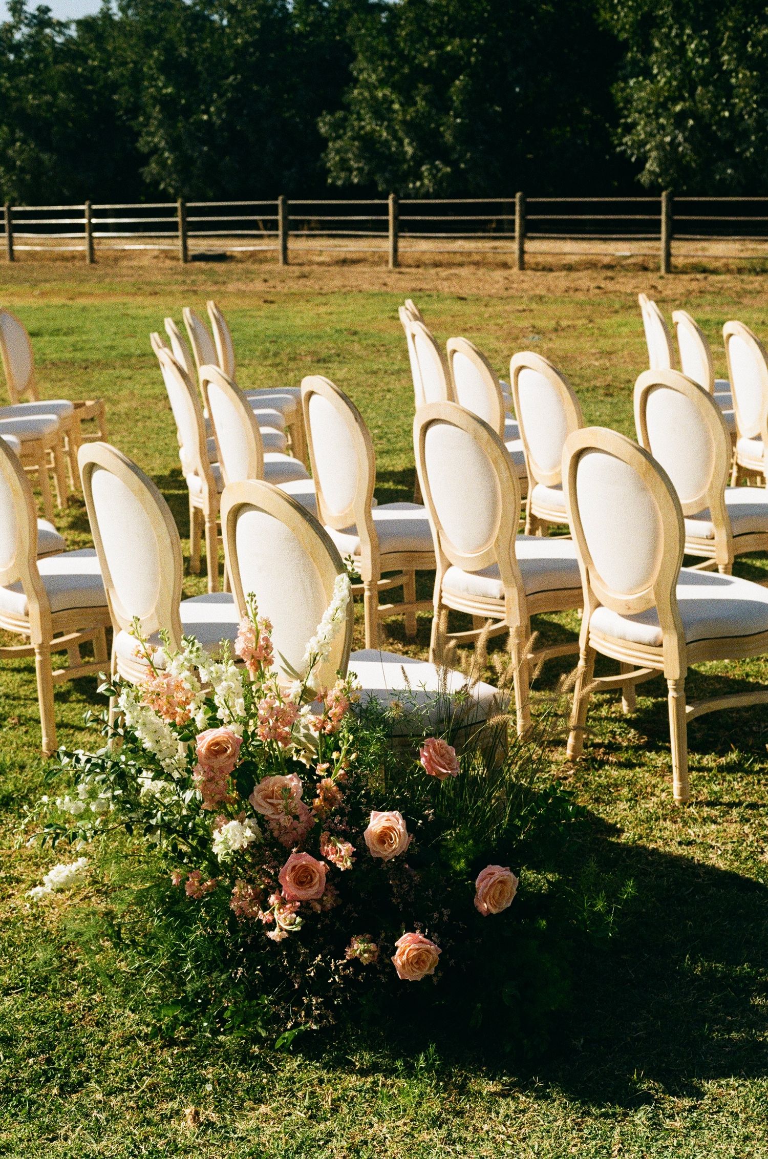 Elegant outdoor wedding ceremony setup with ivory Louis chairs arranged in rows on green grass, adorned with blush pink floral arrangements.