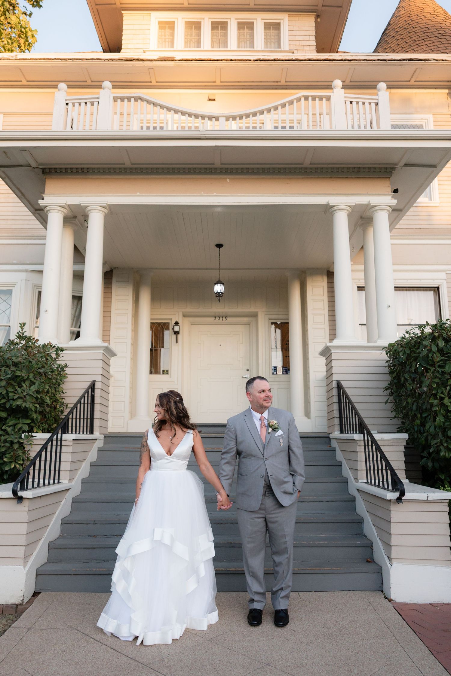 Bride and groom holding hands on steps of elegant Victorian wedding venue at golden hour.