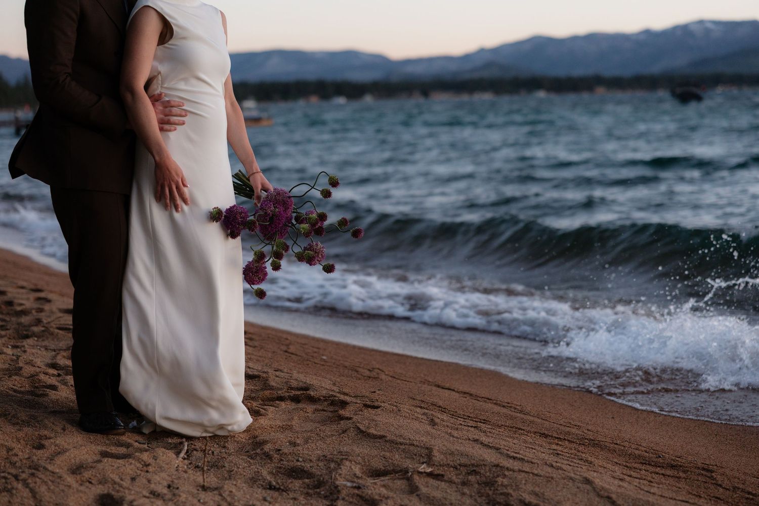 Couple in wedding attire standing on sandy beach at sunset, bride holding purple bouquet with ocean waves behind them.