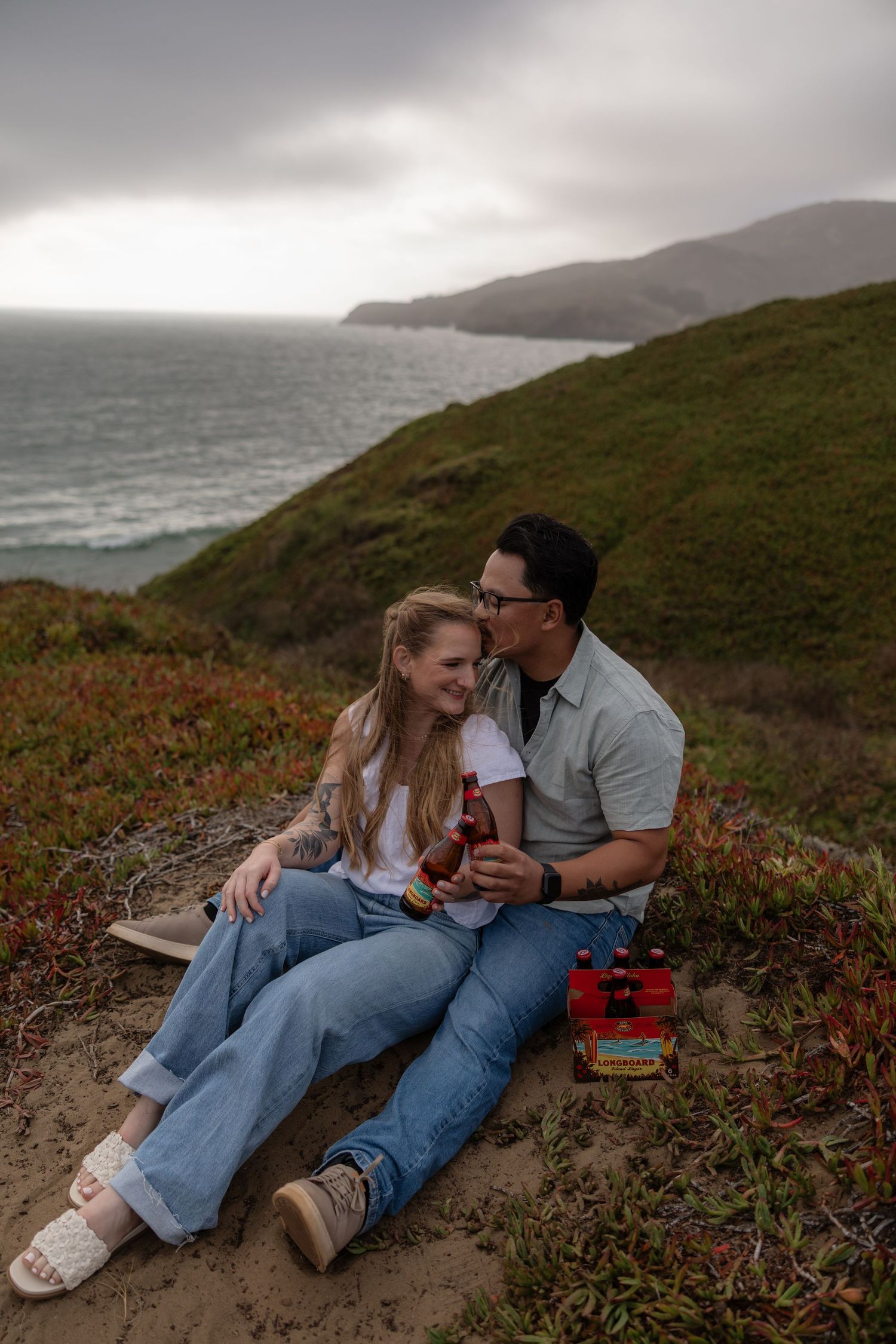 Couple sitting on a coastal hillside, sharing drinks with ocean and green cliffs in the background.