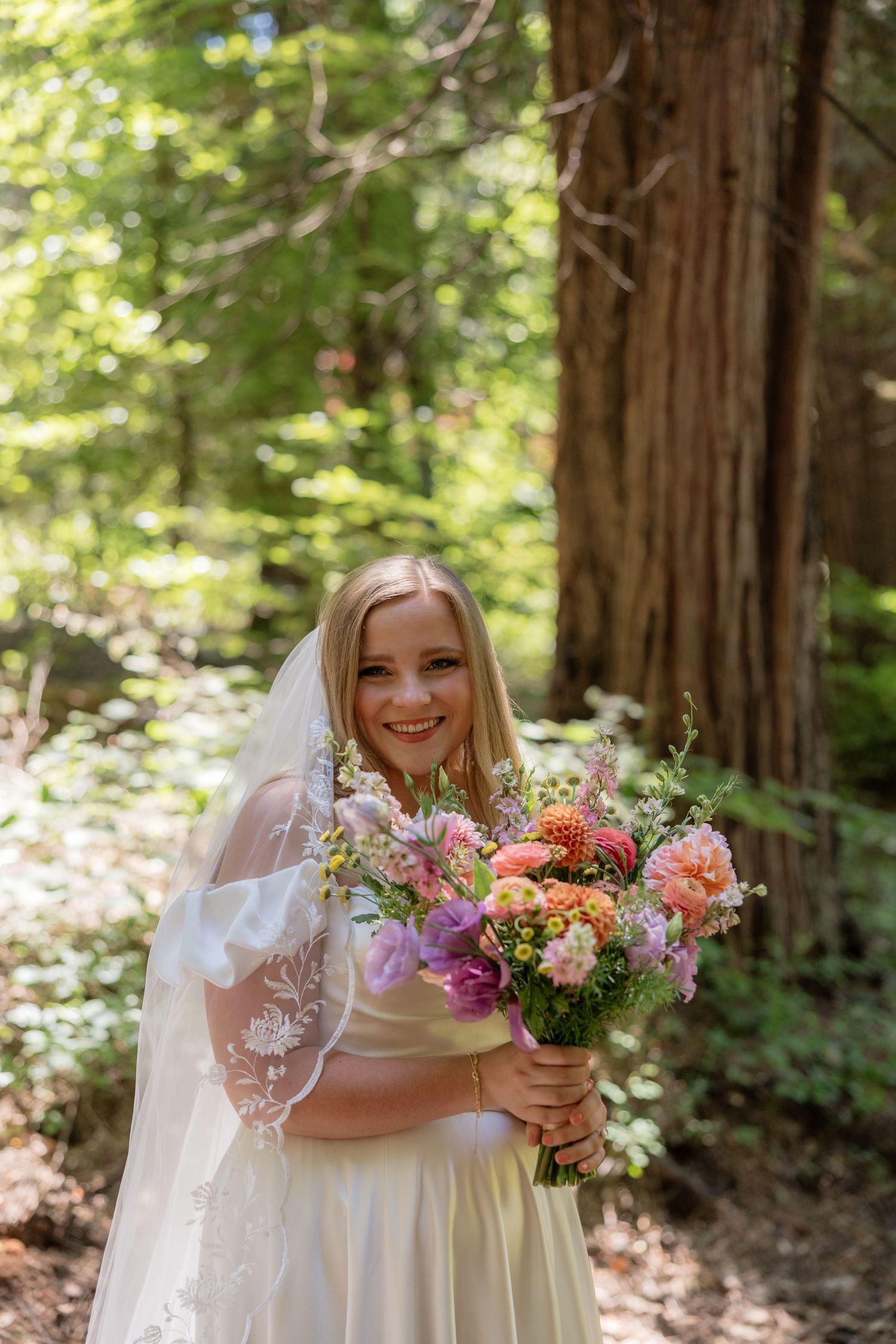 Smiling bride in white lace veil holding colorful bouquet of pink and orange flowers in a sunlit forest.
