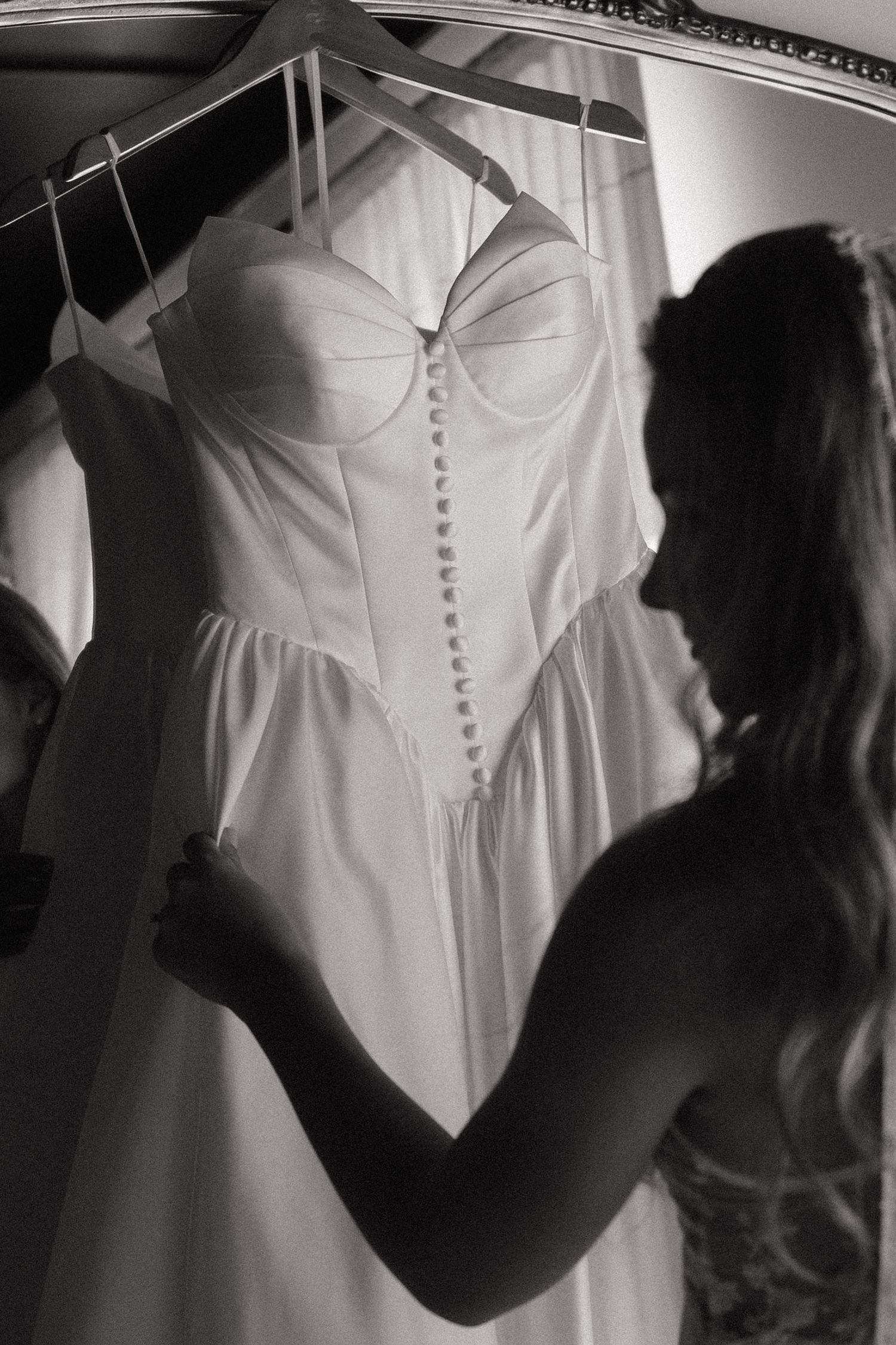 Black and white photo of a bride admiring her elegant corset wedding dress hanging on a rack.