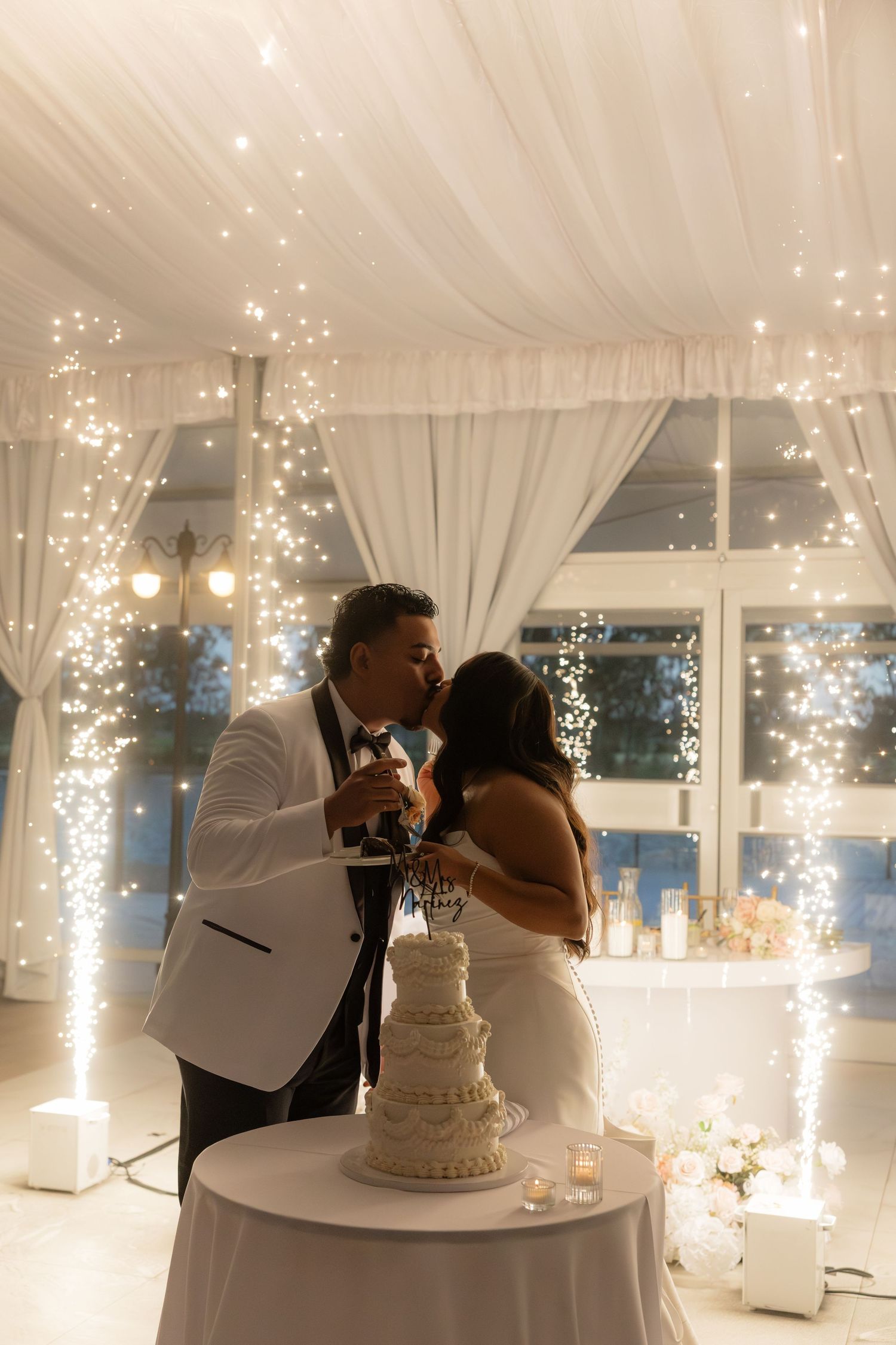 Bride and groom share a kiss while cutting their wedding cake surrounded by sparkling fairy light columns.