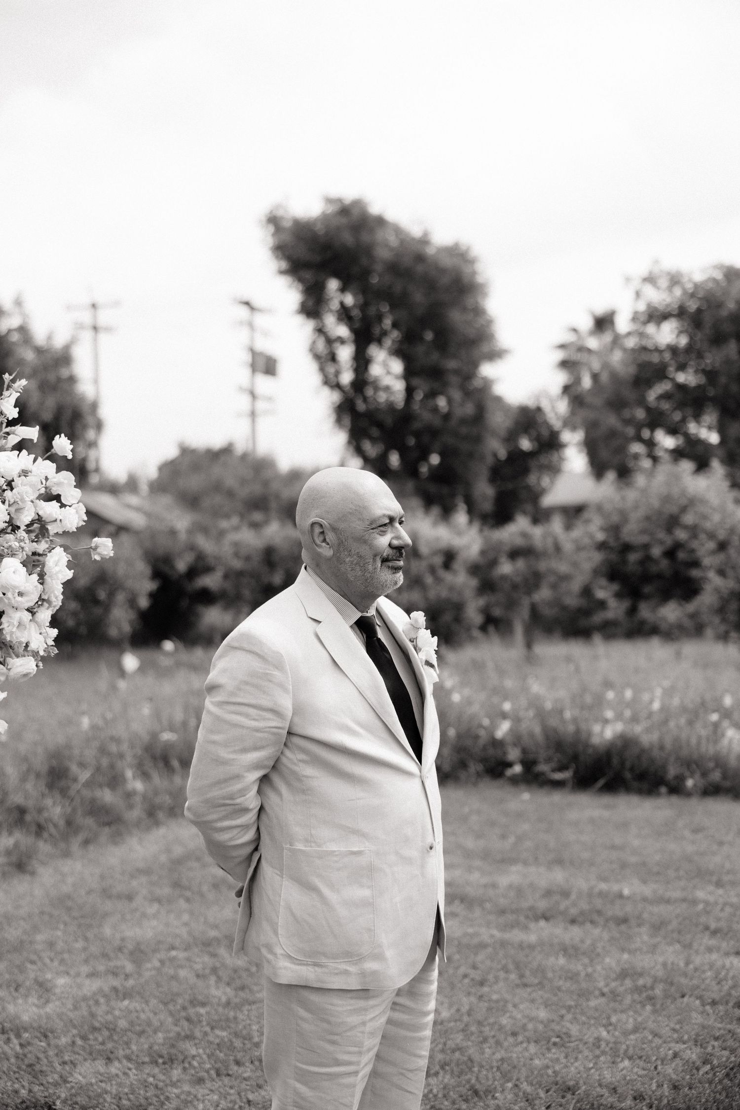 Black and white portrait of a bald groom in a light suit smiling outdoors near blooming flowers.