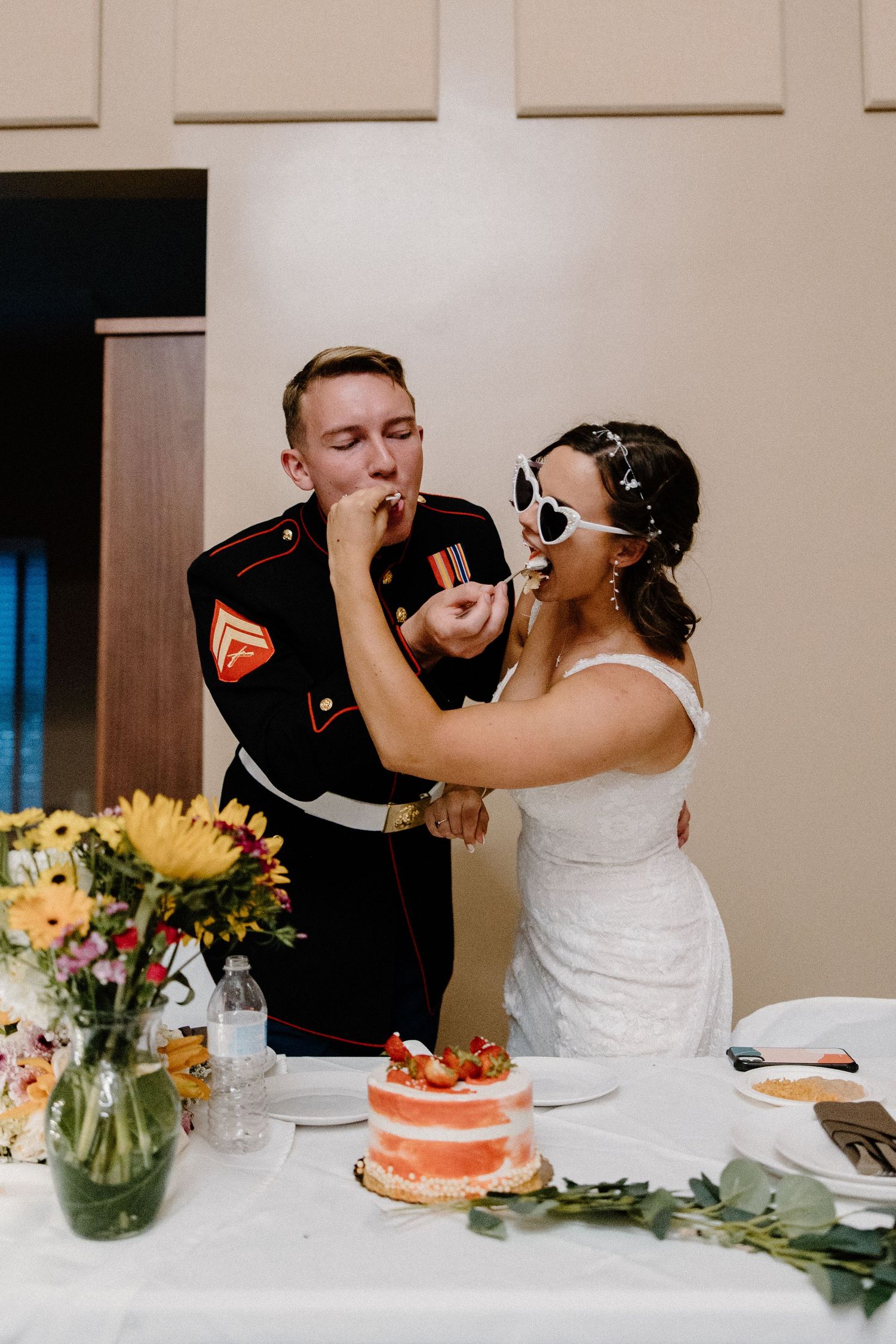 Bride feeds groom wedding cake while wearing heart sunglasses at casual military wedding reception.