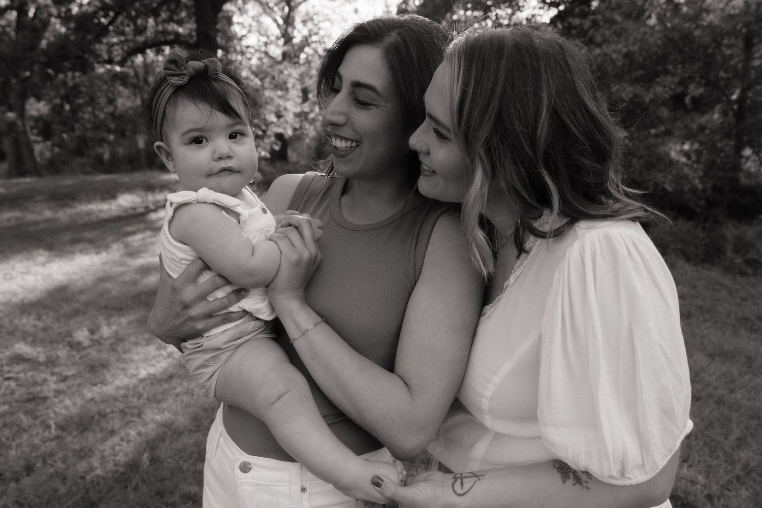 Two women smiling and holding a baby in a black and white outdoor portrait.
