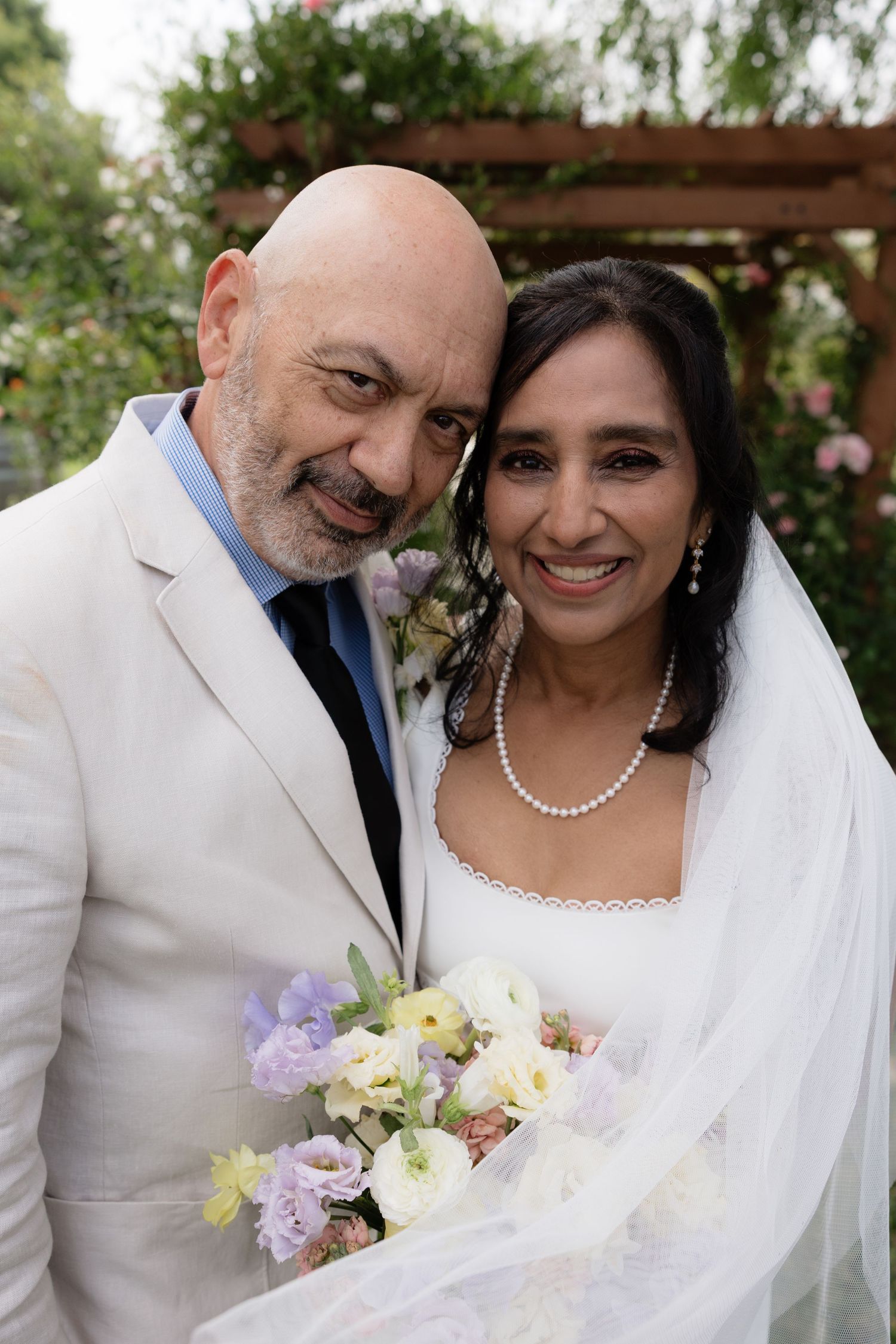 Smiling bride and groom portrait outdoors, bride holding pastel bouquet with white veil, groom in cream suit with boutonniere.