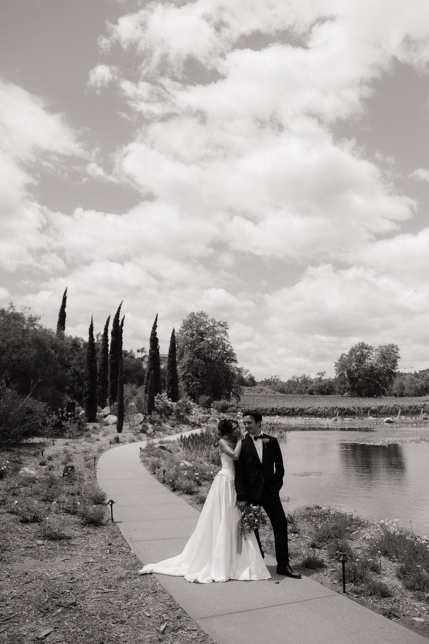 Bride and groom sharing a romantic moment on a lakeside pathway surrounded by lush gardens and cypress trees.