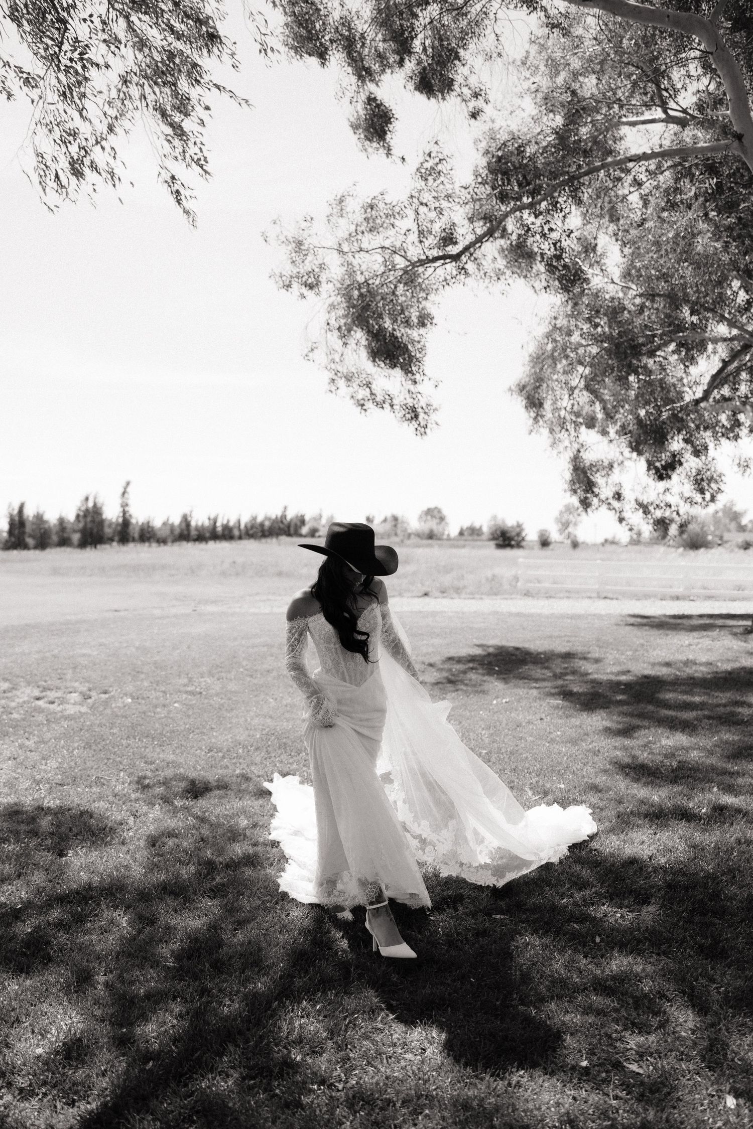 Black and white photo of bride in flowing white dress and black hat twirling under large tree.
