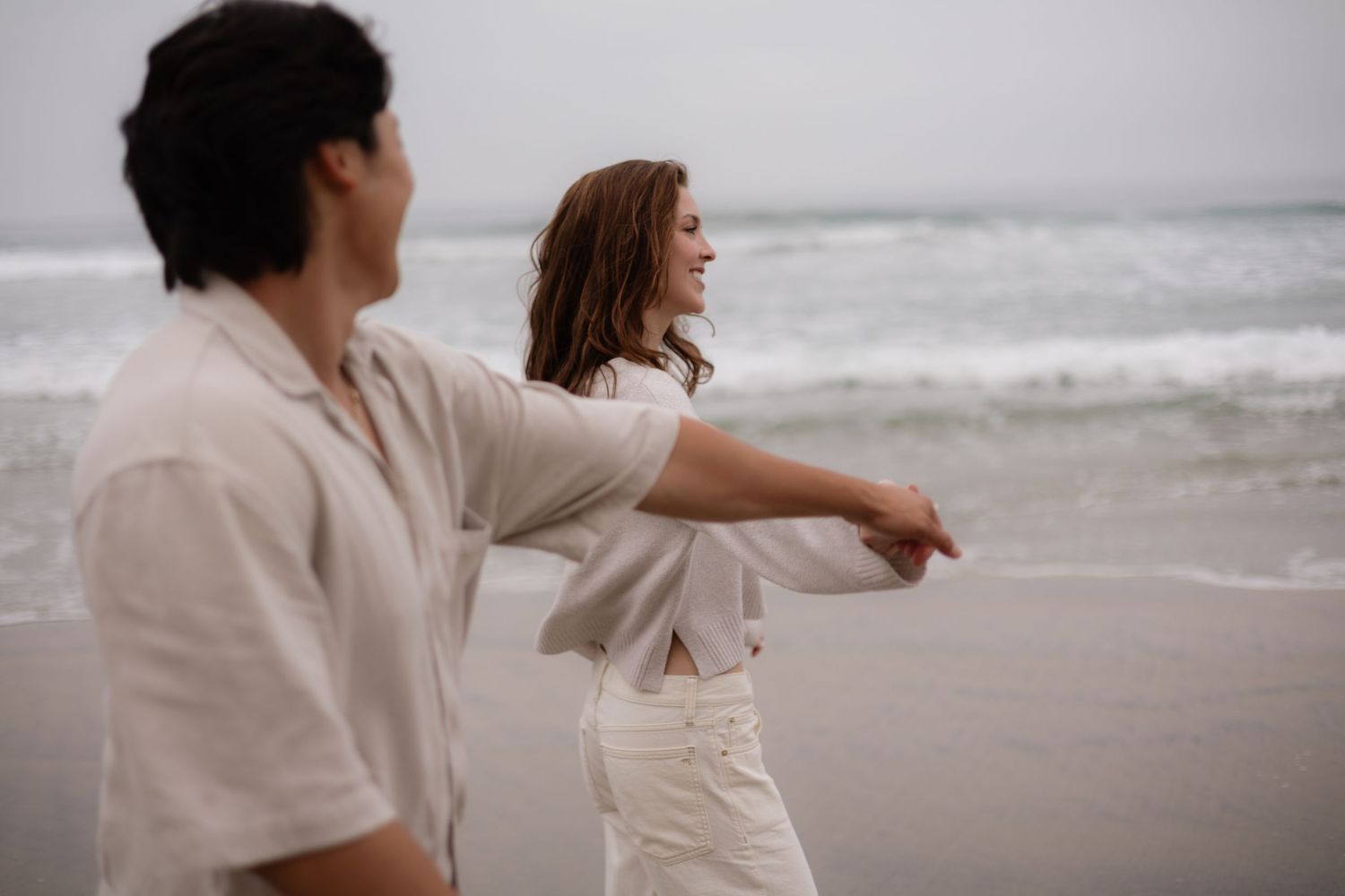 Couple holding hands walking on a beach, woman smiling, wearing white outfits with ocean waves in background.