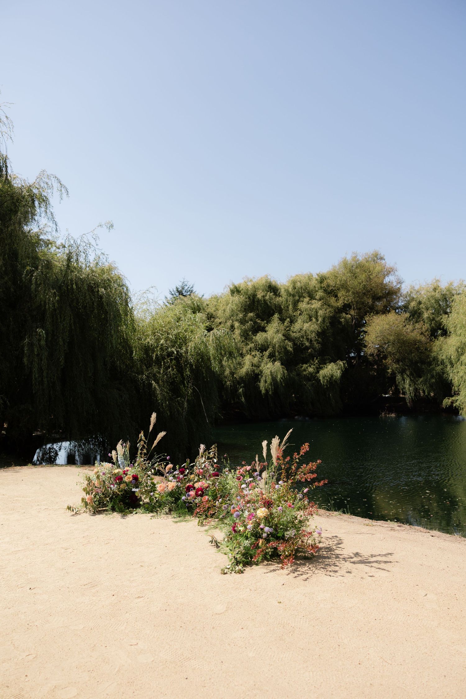 Colorful wildflower arrangement on sandy shore beside a tranquil green lake surrounded by weeping willow trees.