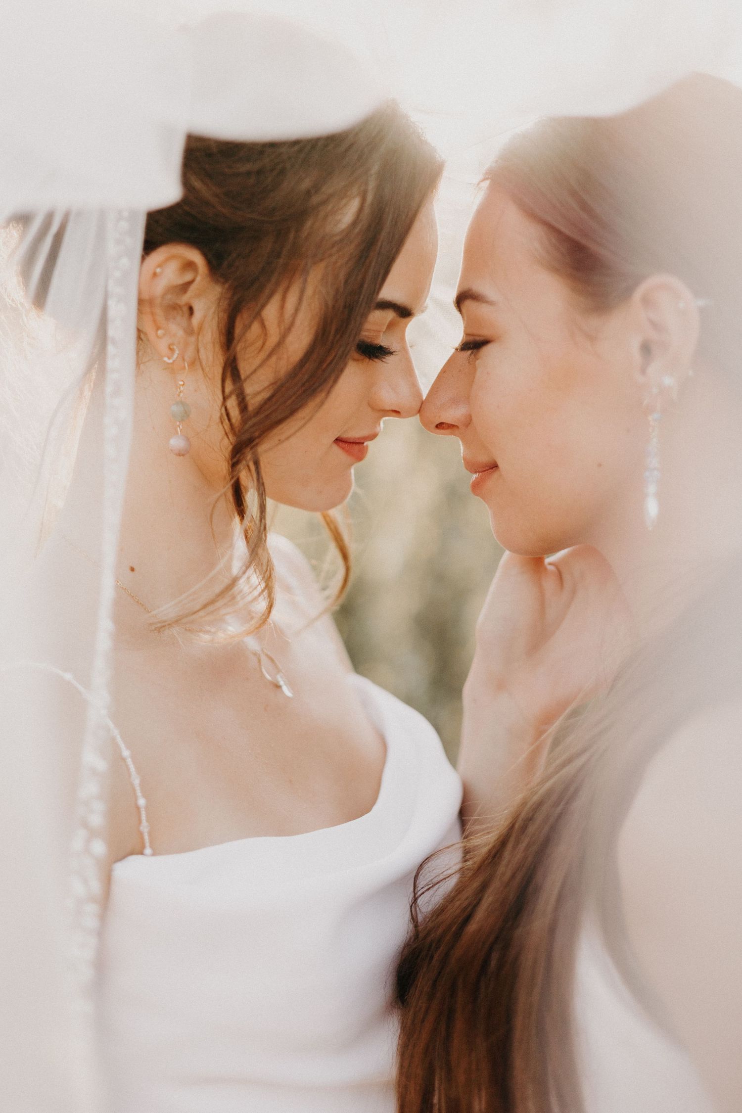 Two brides sharing an intimate forehead-to-forehead moment beneath a flowing veil in soft sunlight.