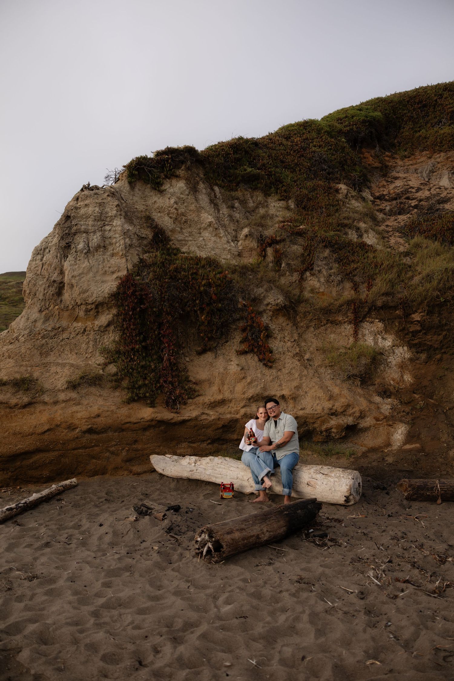 Couple sitting on driftwood logs on a sandy beach with dramatic rocky cliffs behind them.