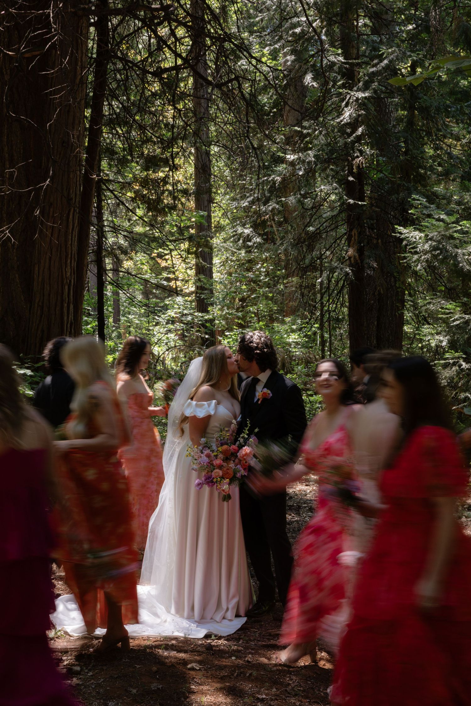 A bride and groom share a first kiss surrounded by wedding guests in a lush forest setting.